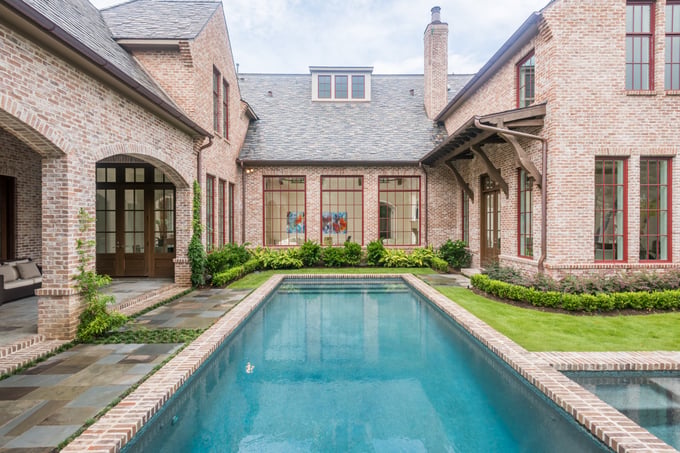 Sims Builders Houston French-inspired brick custom home with red-trimmed steel windows, brick-edged rectangular lap pool, and slate courtyard