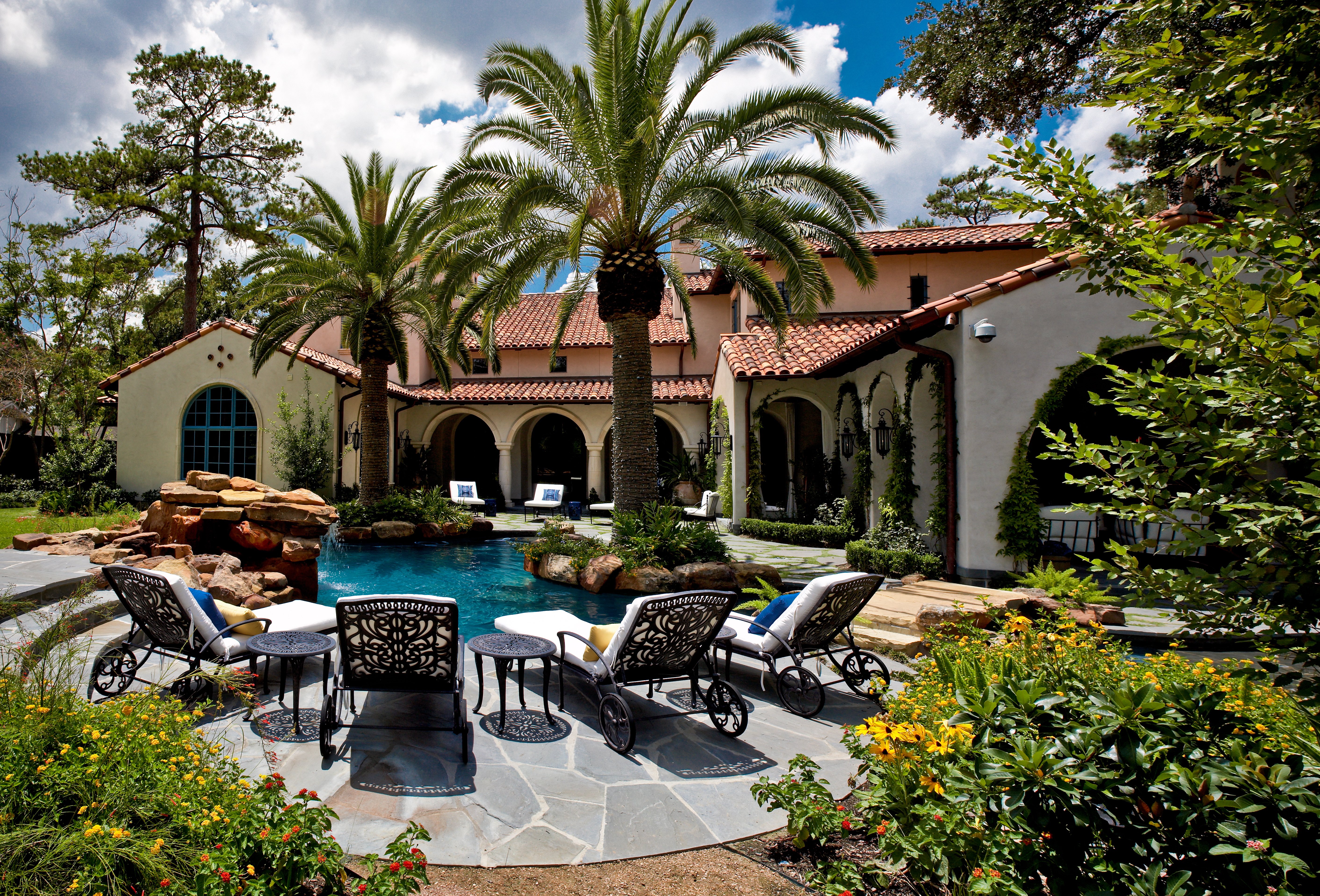 Sims Builders Houston Spanish Mediterranean custom home with arched loggia, palm trees, cast iron lounge chairs, and natural stone pool patio