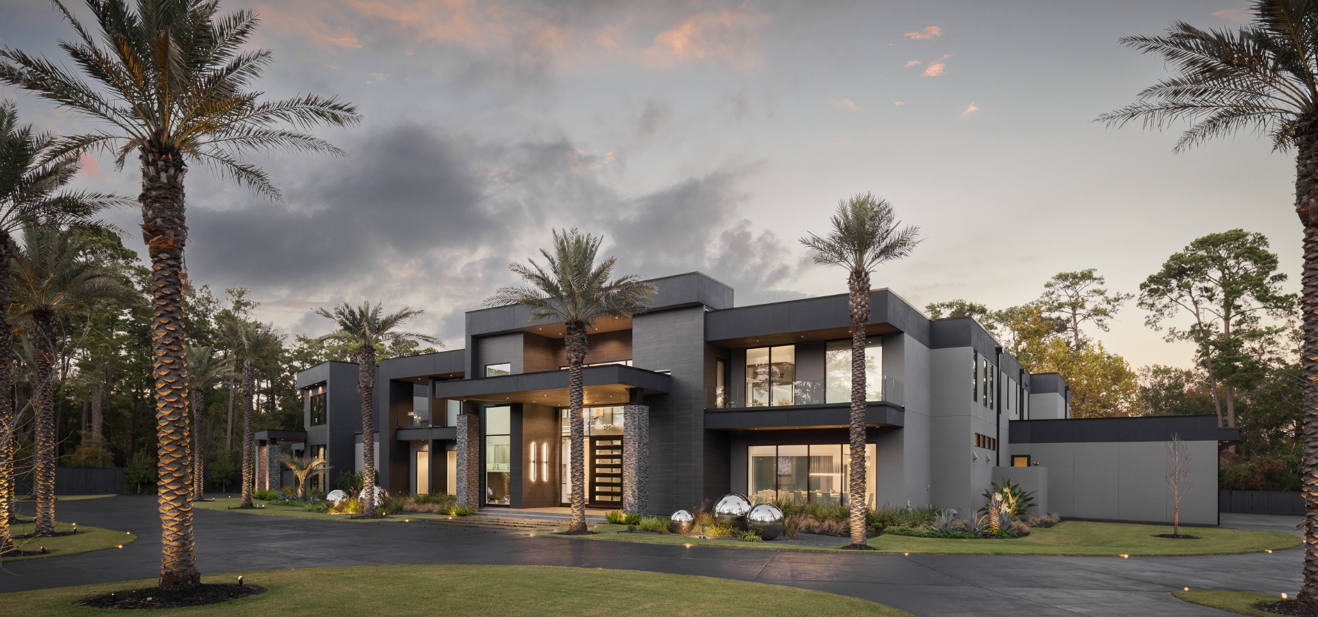 Contemporary luxury custom home in Houston with dramatic modern architecture, palm-lined driveway, dark stone and stucco exterior, expansive windows, and warm entry lighting at dusk creating a striking modern estate presence. Contemporary luxury custom home in Houston with dramatic modern architecture, palm-lined driveway, dark stone and stucco exterior, expansive windows, and warm entry lighting at dusk creating a striking modern estate presence.