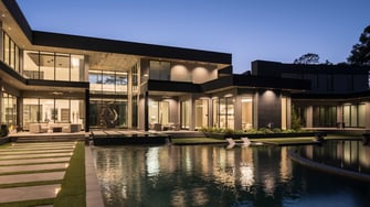 Modern luxury Houston custom home at dusk featuring expansive glass walls, two-story architecture, resort-style pool with sun shelves, outdoor living terraces, and dramatic contemporary lighting reflecting across the water.