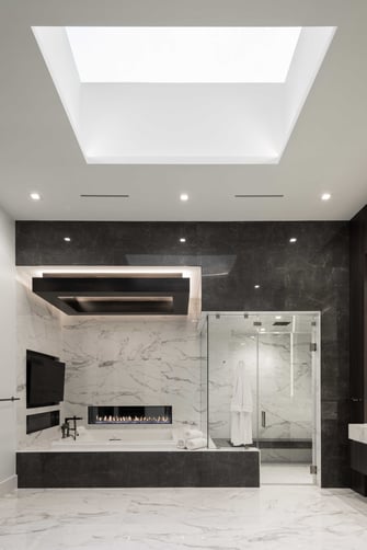 Luxury spa bathroom in a modern custom home featuring marble walls and floors, freestanding soaking tub with linear fireplace, glass walk-in shower, and large recessed skylight creating a bright contemporary retreat.