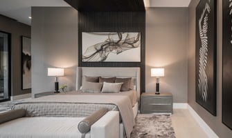 Modern luxury bedroom in a Houston custom home featuring a tall upholstered bed, dark wood accent wall, abstract artwork, matching bedside tables with lamps, neutral tones, and layered textures creating a calm contemporary retreat.