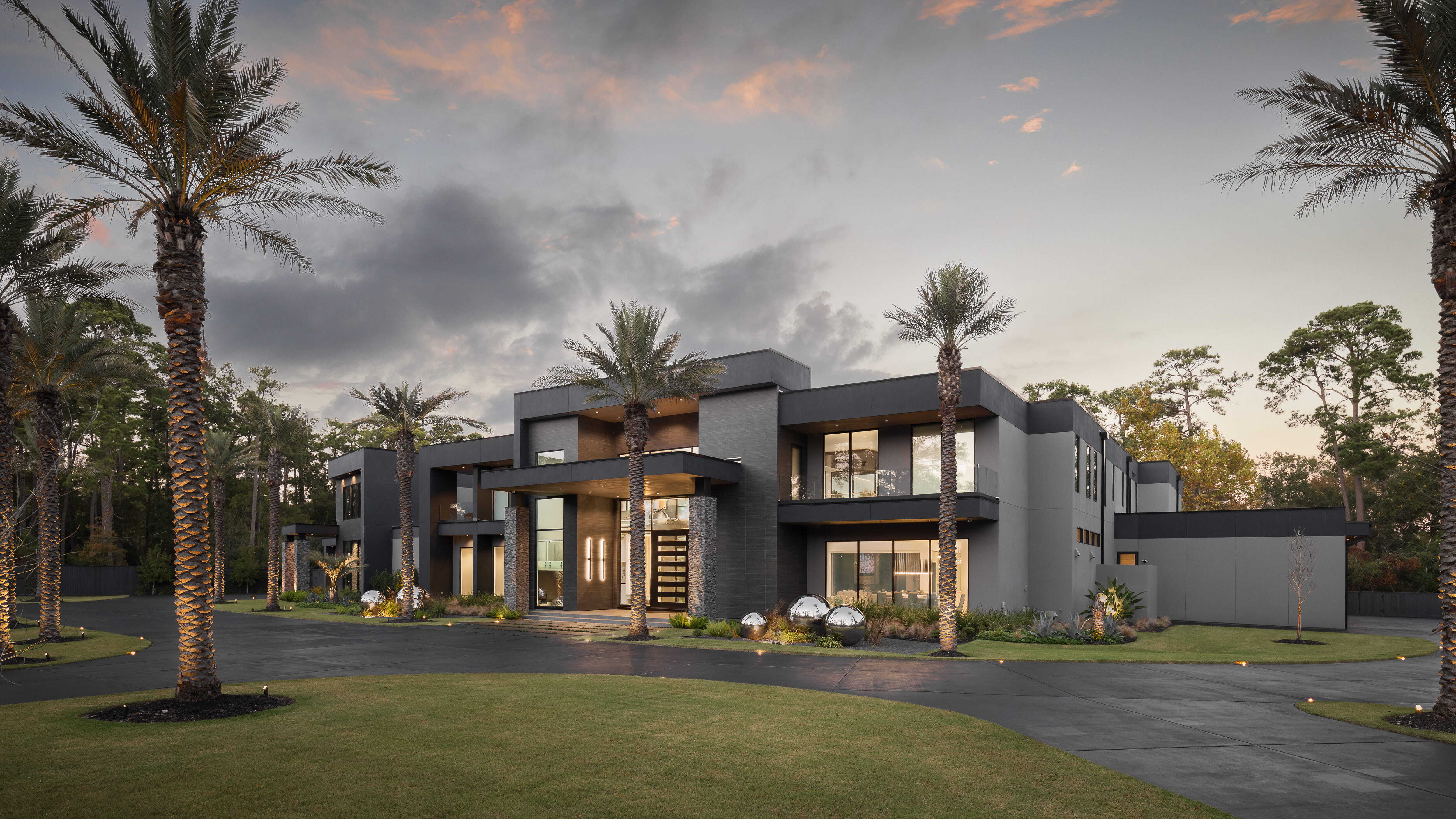 Modern luxury custom home exterior in Memorial Houston featuring clean architectural lines, expansive glass, palm-lined drive, dramatic lighting, and landscaped grounds at sunset.
