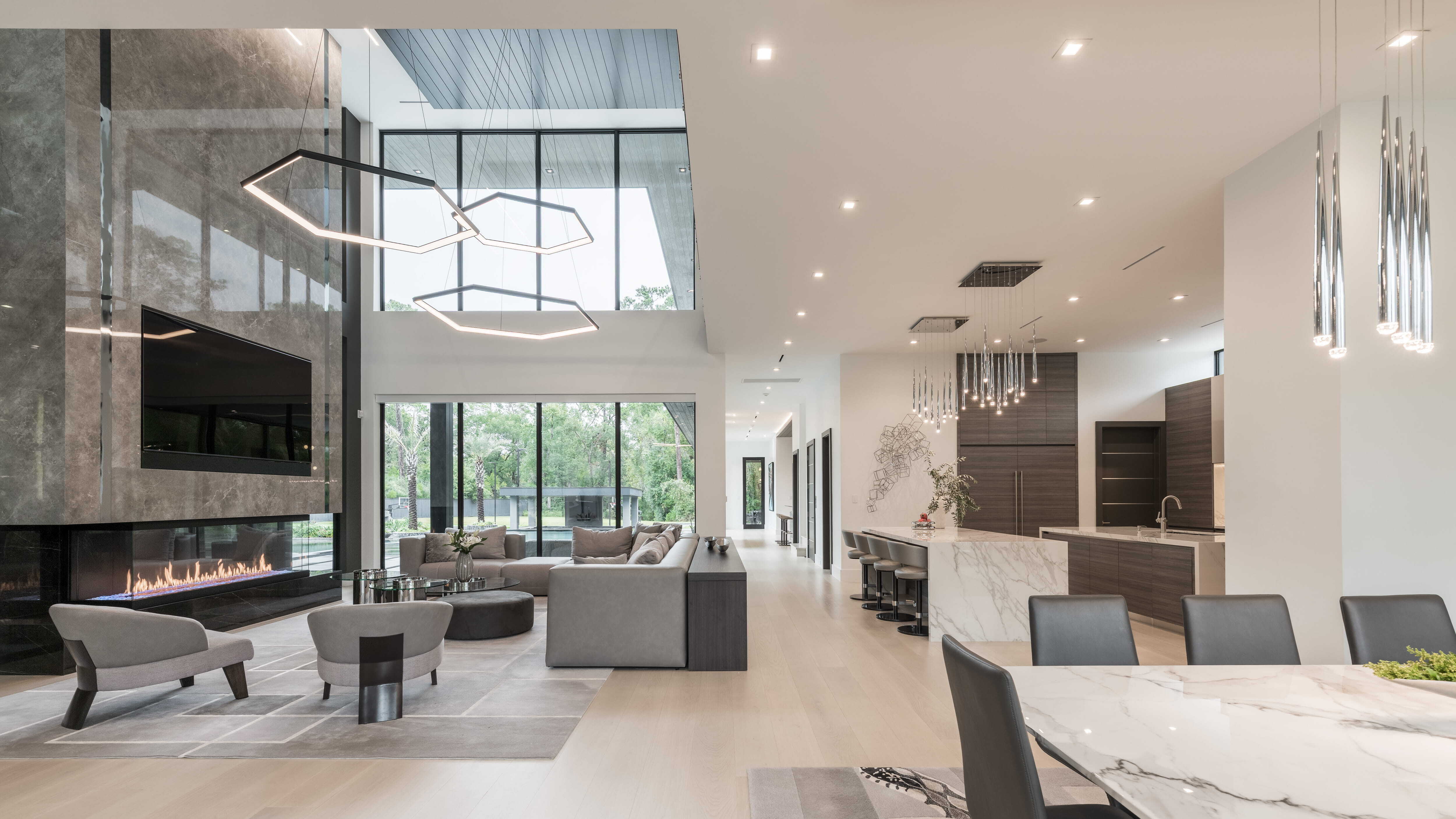 Open living room and kitchen with floor-to-ceiling windows, marble waterfall island, contemporary lighting, modern fireplace, and indoor-outdoor views in a luxury modern Houston custom home.