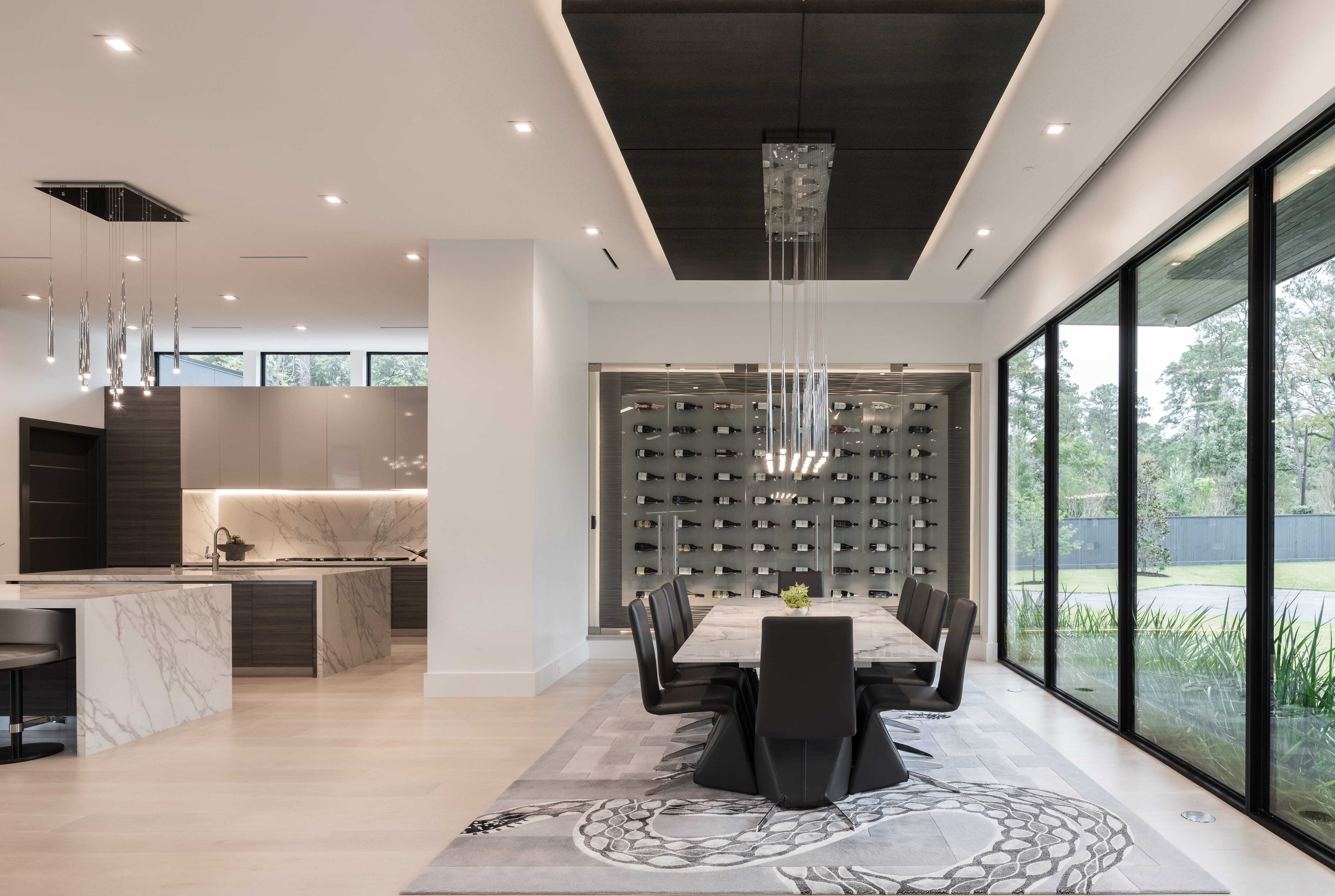 Modern dining room with marble table, sculptural pendant lighting, and a glass wine wall connecting to a sleek contemporary kitchen with waterfall island in a luxury Memorial Houston custom home.