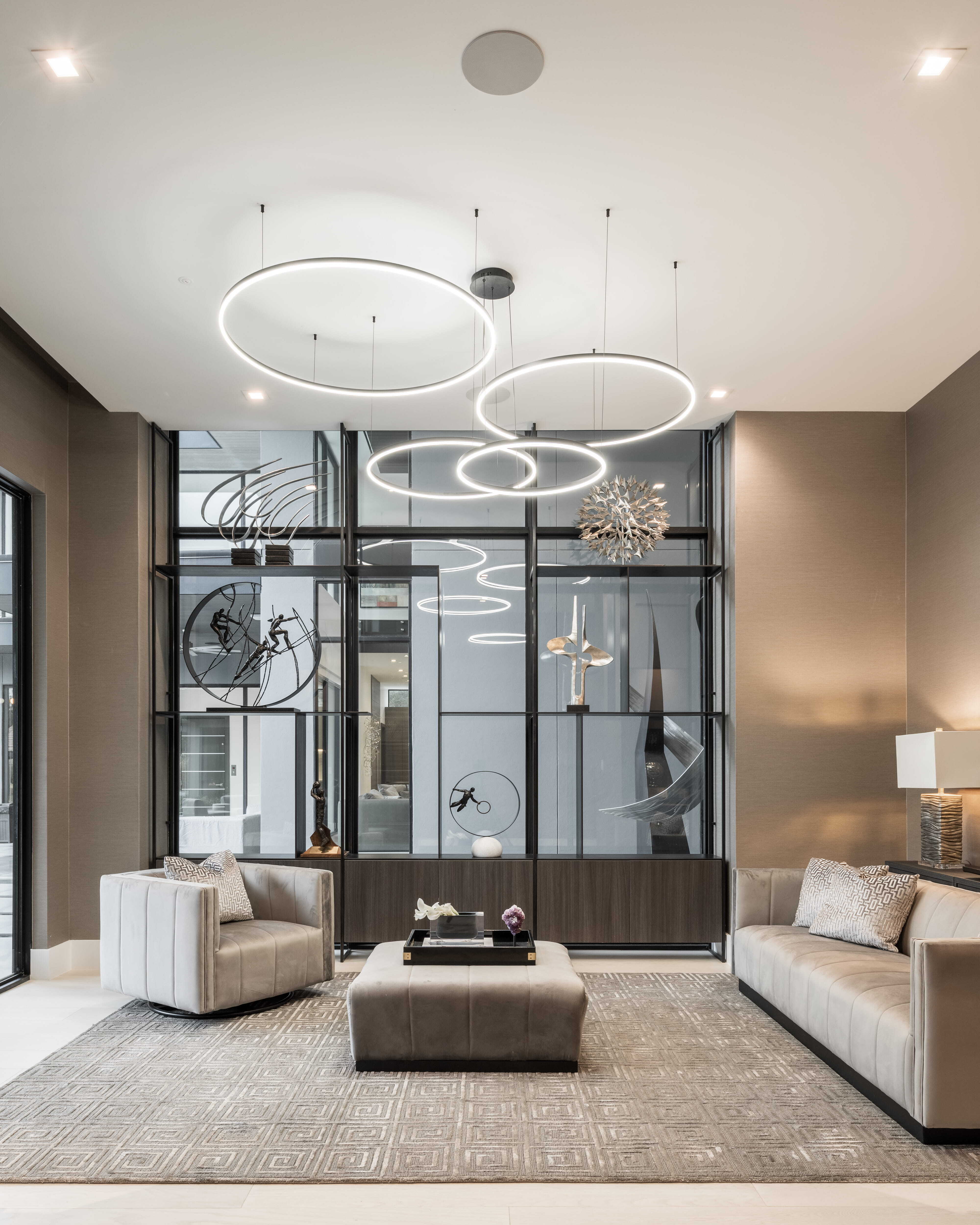 Contemporary sitting room with sculptural ring chandeliers, neutral seating, and a glass display wall showcasing modern art and sculpture inside a luxury custom home in Memorial Houston.