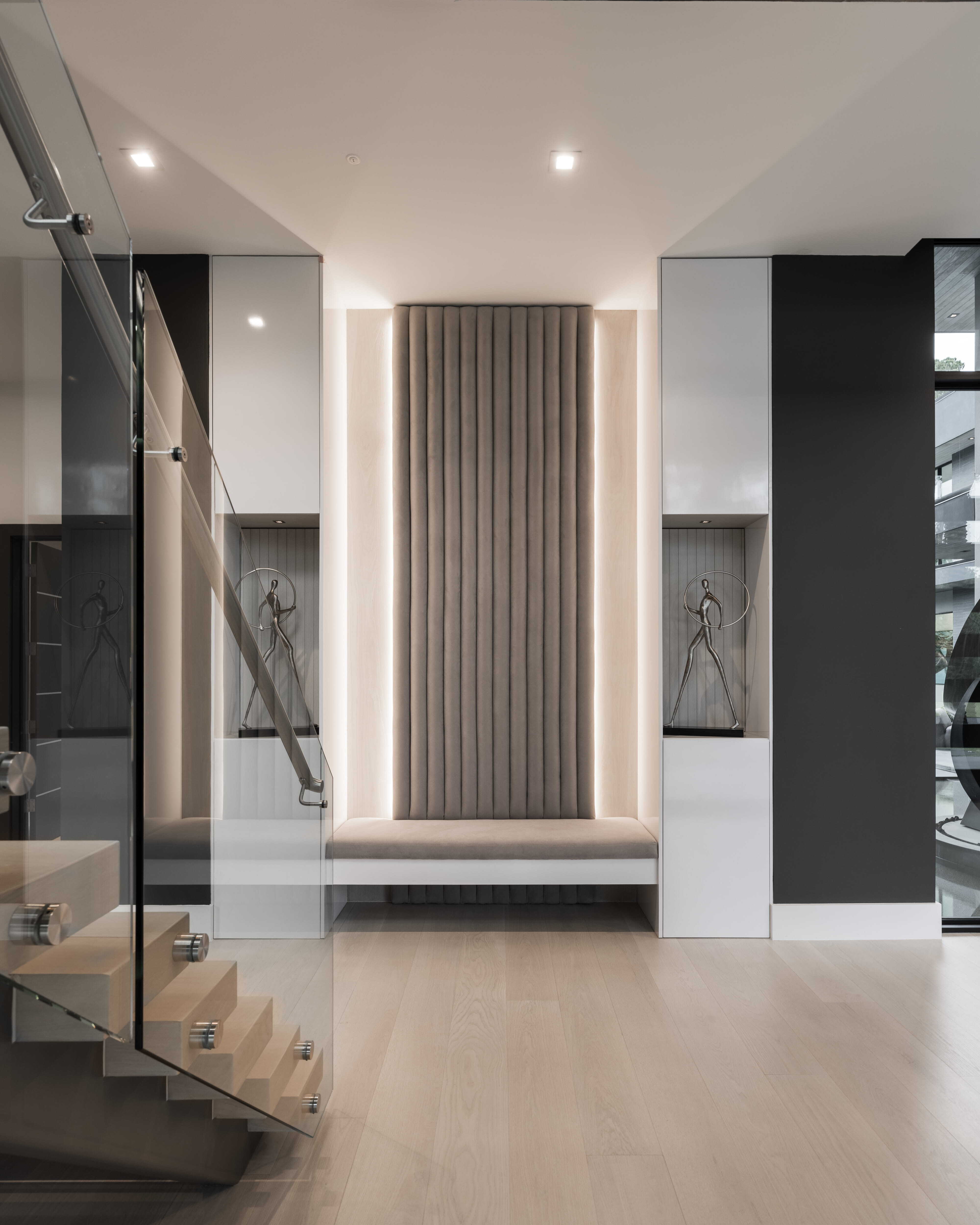 Modern stair hall with floating wood staircase, glass railing, and upholstered built-in bench framed by soft lighting and sculptural display niches inside a luxury Memorial Houston custom home.