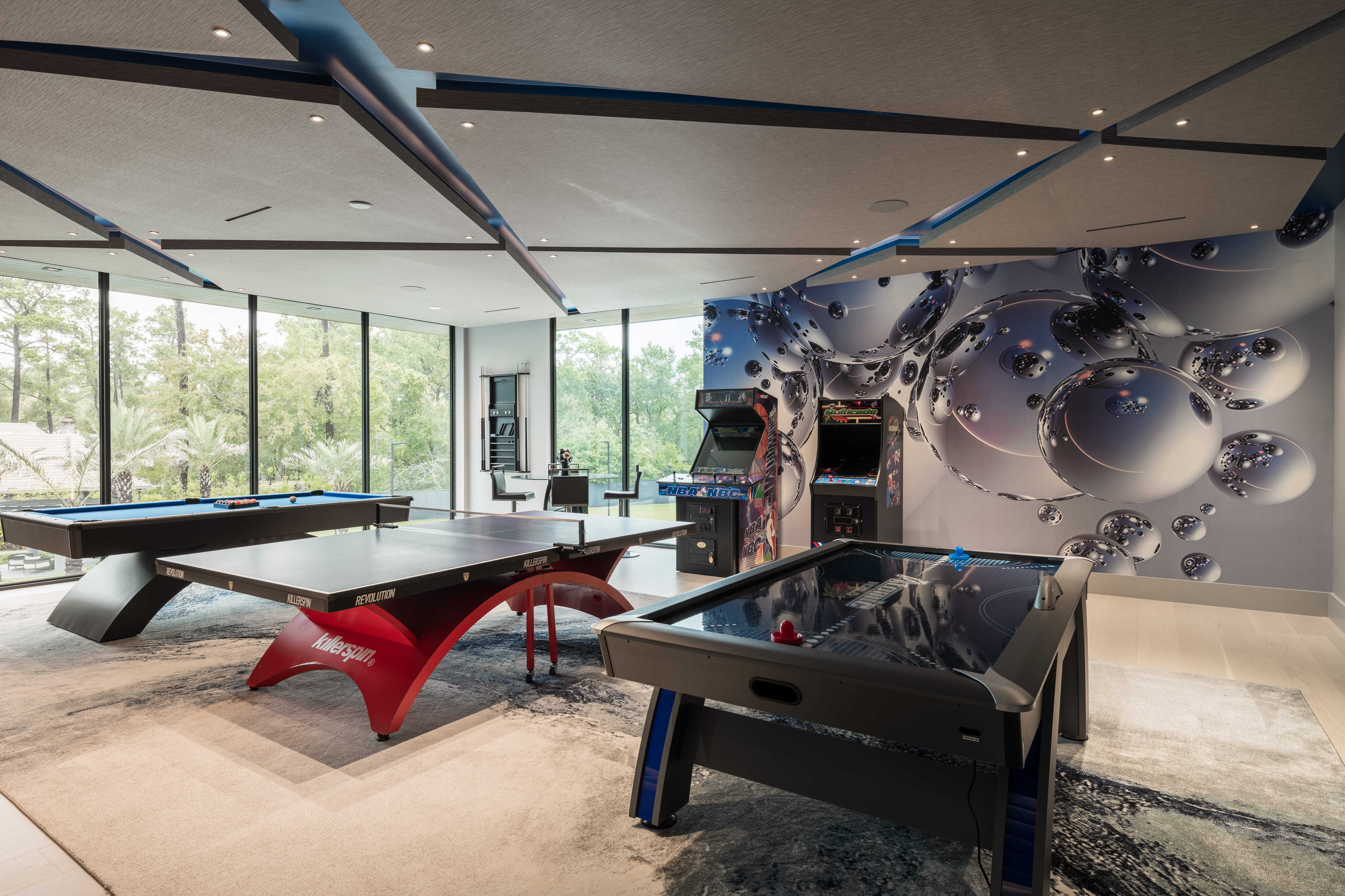 Luxury game room with ping pong table, pool table, air hockey, and classic arcade games surrounded by floor-to-ceiling windows overlooking the landscaped grounds of a modern custom home.