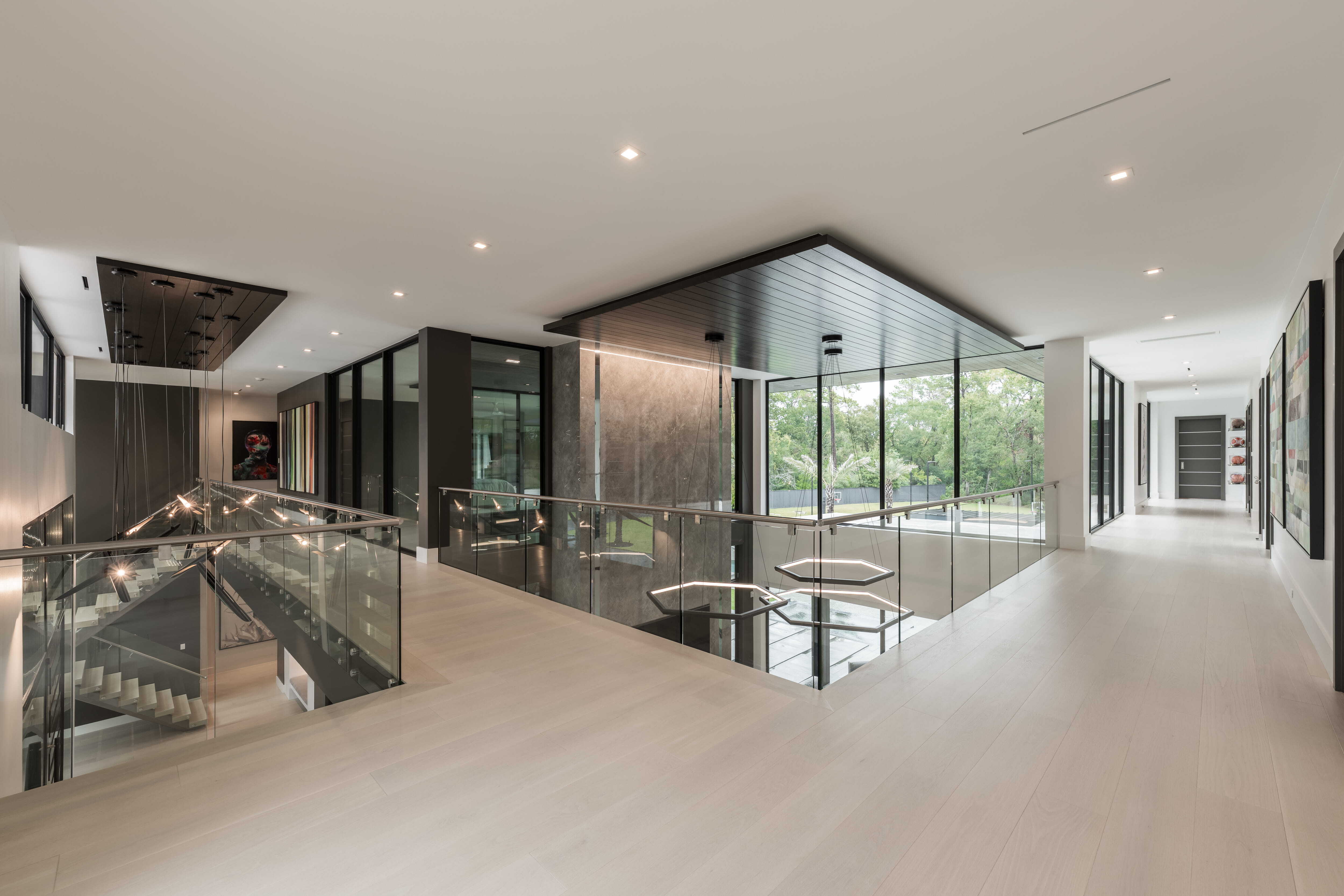 Second-floor gallery hallway overlooking a dramatic two-story living space with glass railings, sculptural lighting, and floor-to-ceiling windows in a modern luxury custom home in Memorial Houston.