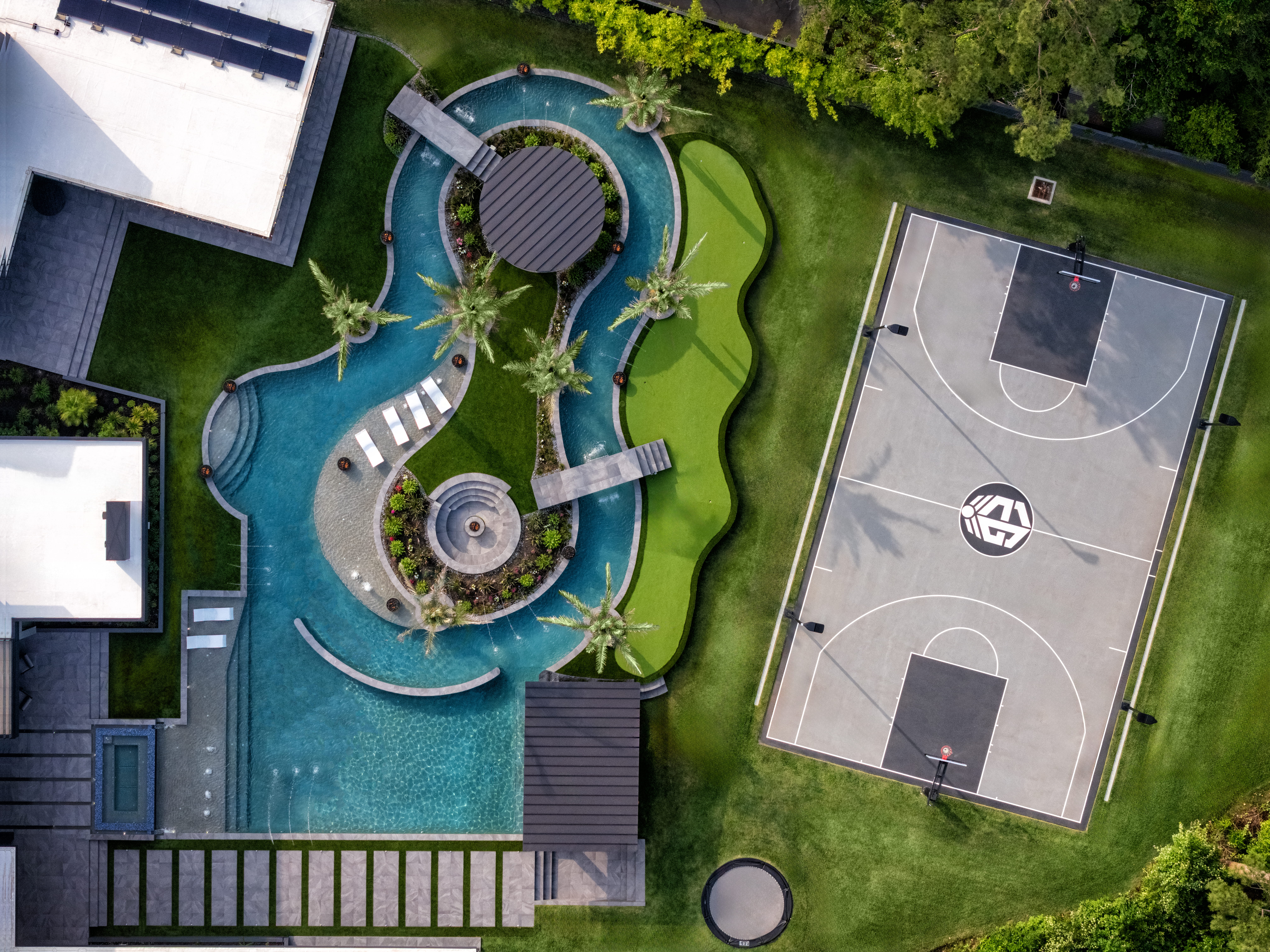 Aerial view of a modern luxury estate in Memorial Houston featuring a resort-style pool with lazy river, spa islands, palm trees, putting green, and a private basketball court beside the contemporary custom home.