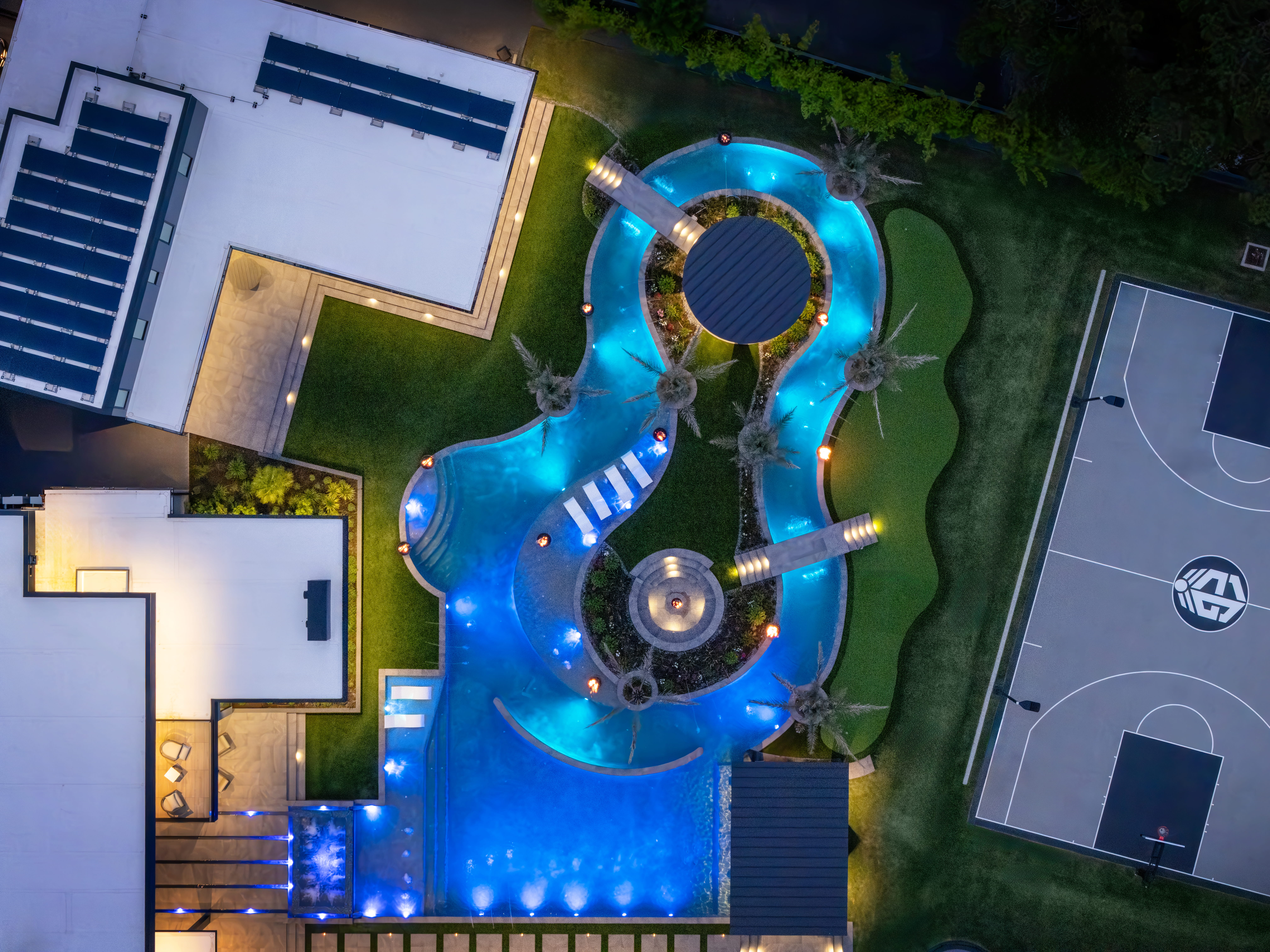 Aerial view of illuminated resort-style pool with lazy river, fire features, and swim-up island beside a private basketball court at a modern luxury estate in Memorial Houston.