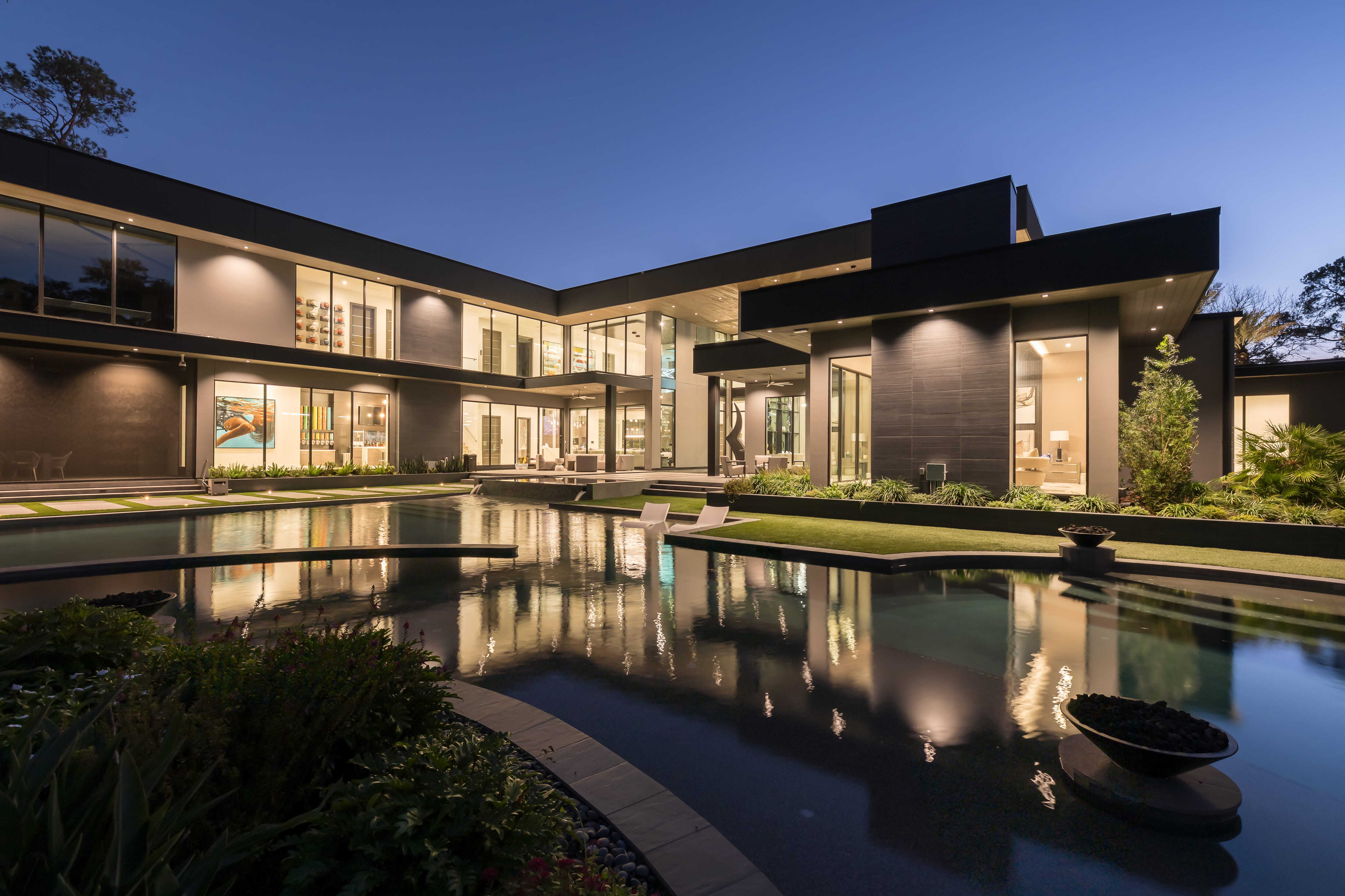 Rear exterior of a modern luxury custom home in Memorial Houston with expansive glass walls overlooking a resort-style pool and outdoor living areas illuminated at dusk.