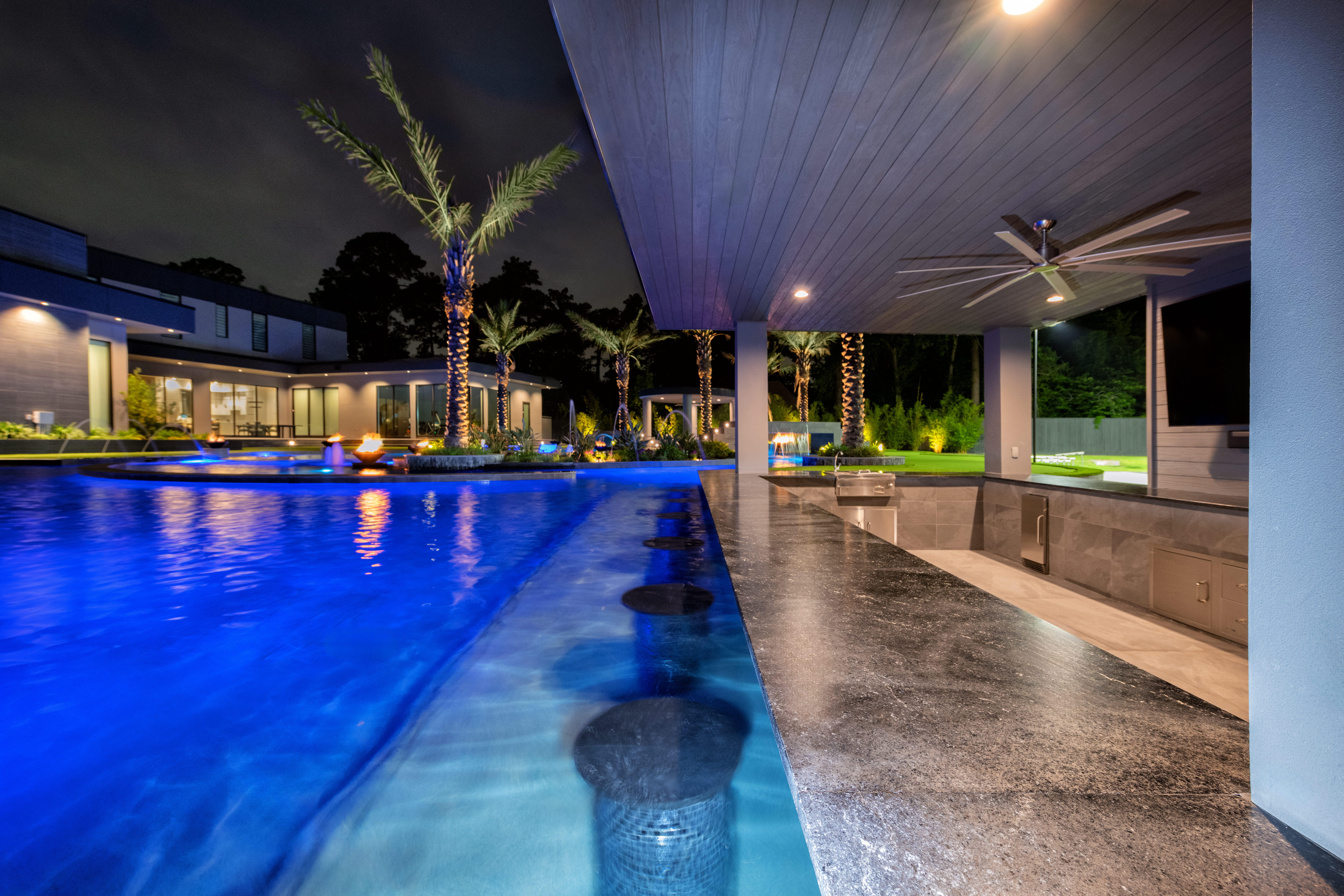 Swim-up bar with submerged seating beside a covered outdoor kitchen and lounge area overlooking an illuminated resort-style pool at a modern luxury custom home in Memorial Houston.