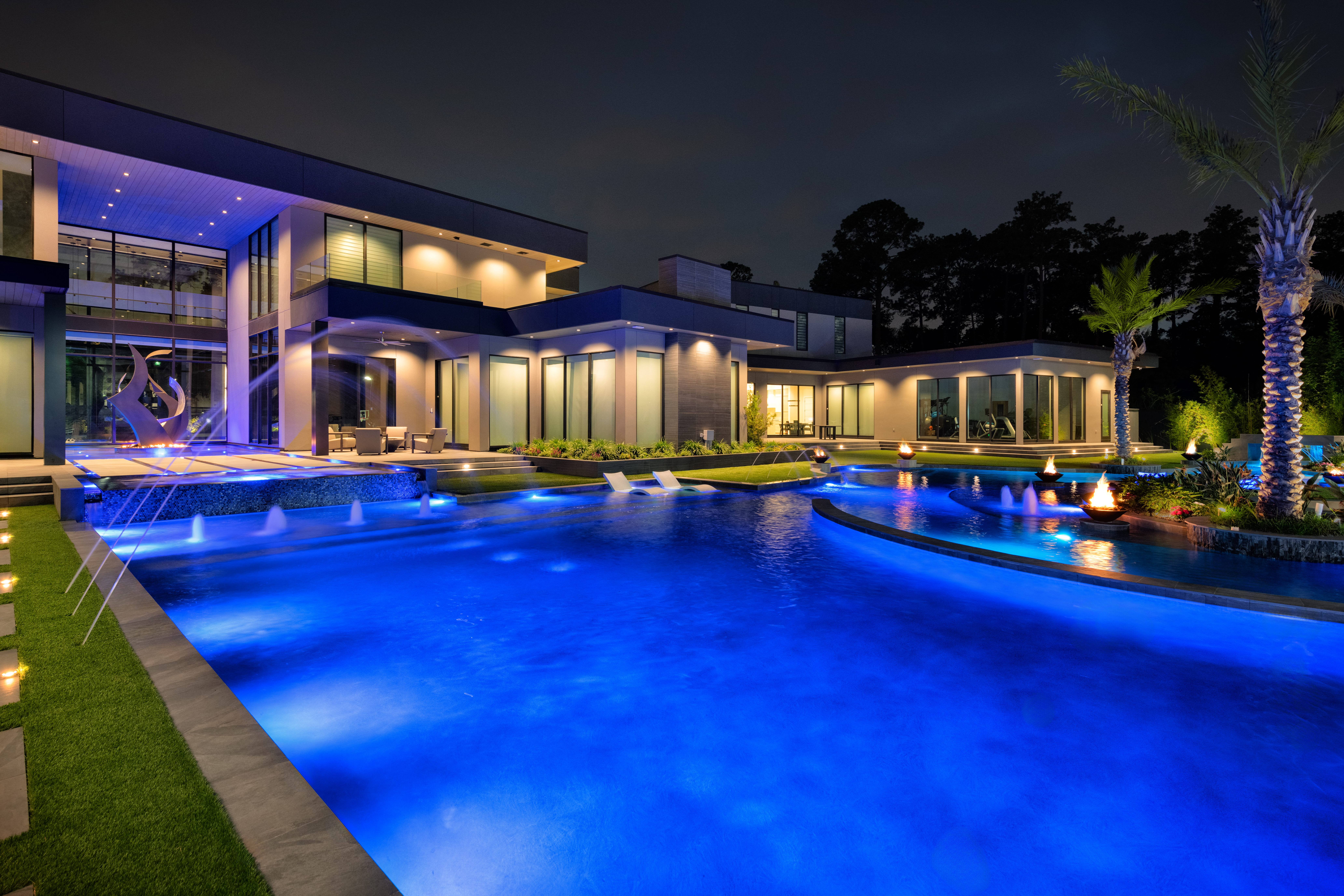 Resort-style backyard pool with raised spa, water fountains, fire bowls, and palm landscaping beside a contemporary luxury custom home in Memorial Houston.