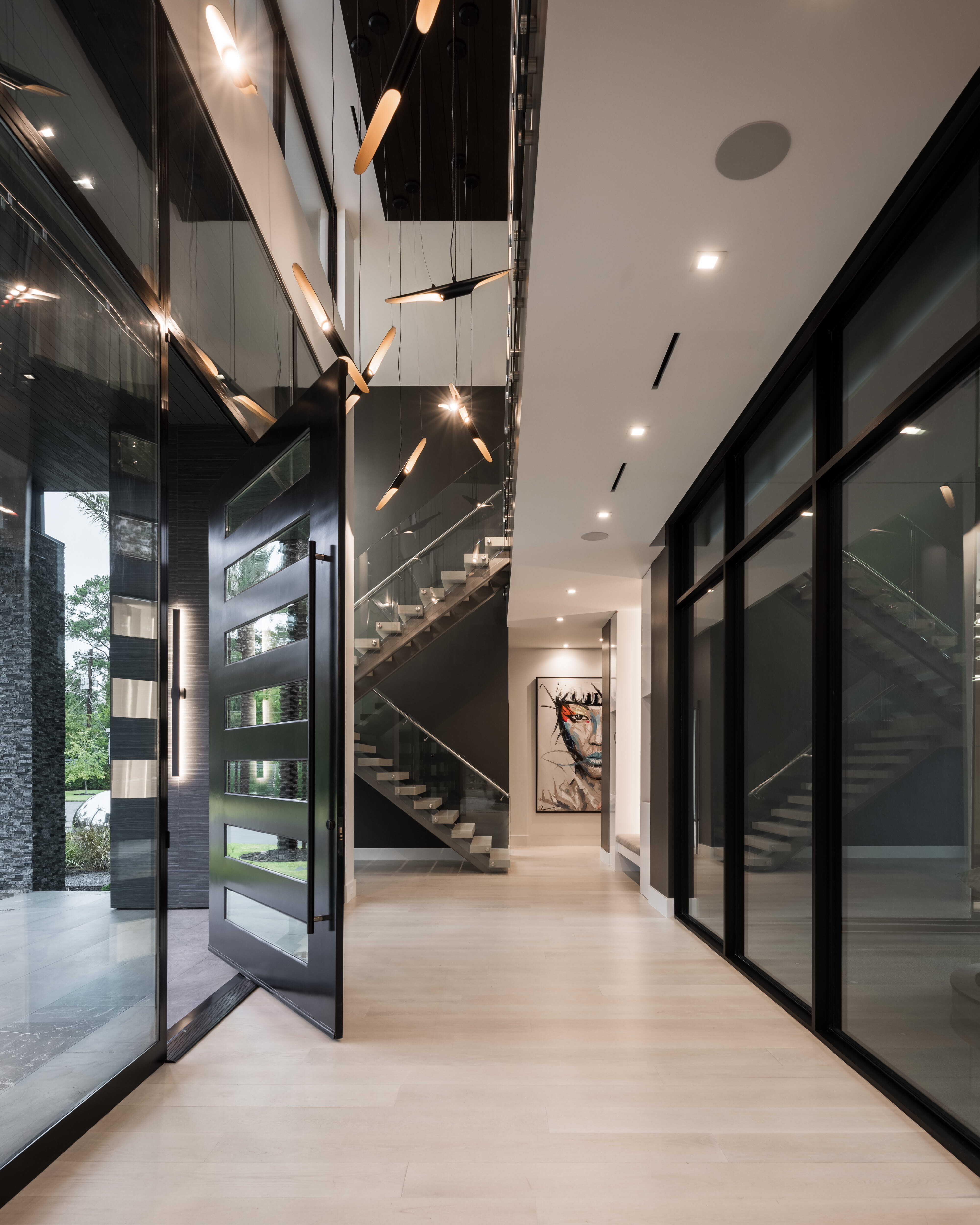 Modern luxury entry with oversized steel pivot front door, floating staircase with glass railing, sculptural pendant lighting, and floor-to-ceiling glass walls creating a dramatic two-story foyer in a contemporary Memorial Houston custom home.
