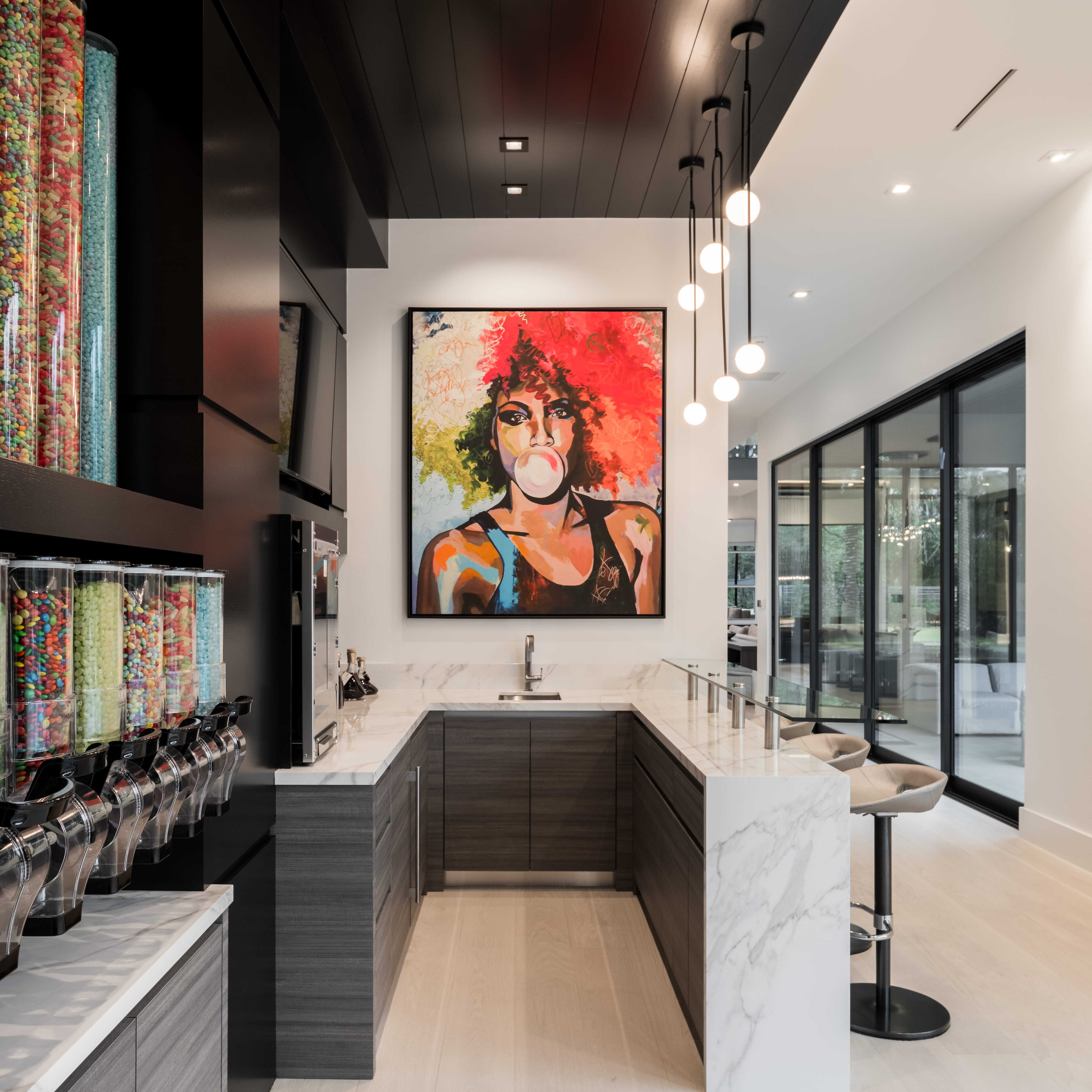 Modern candy bar and beverage station with marble counters, colorful candy dispensers, and bold contemporary artwork creating a playful entertainment space inside a luxury custom home.