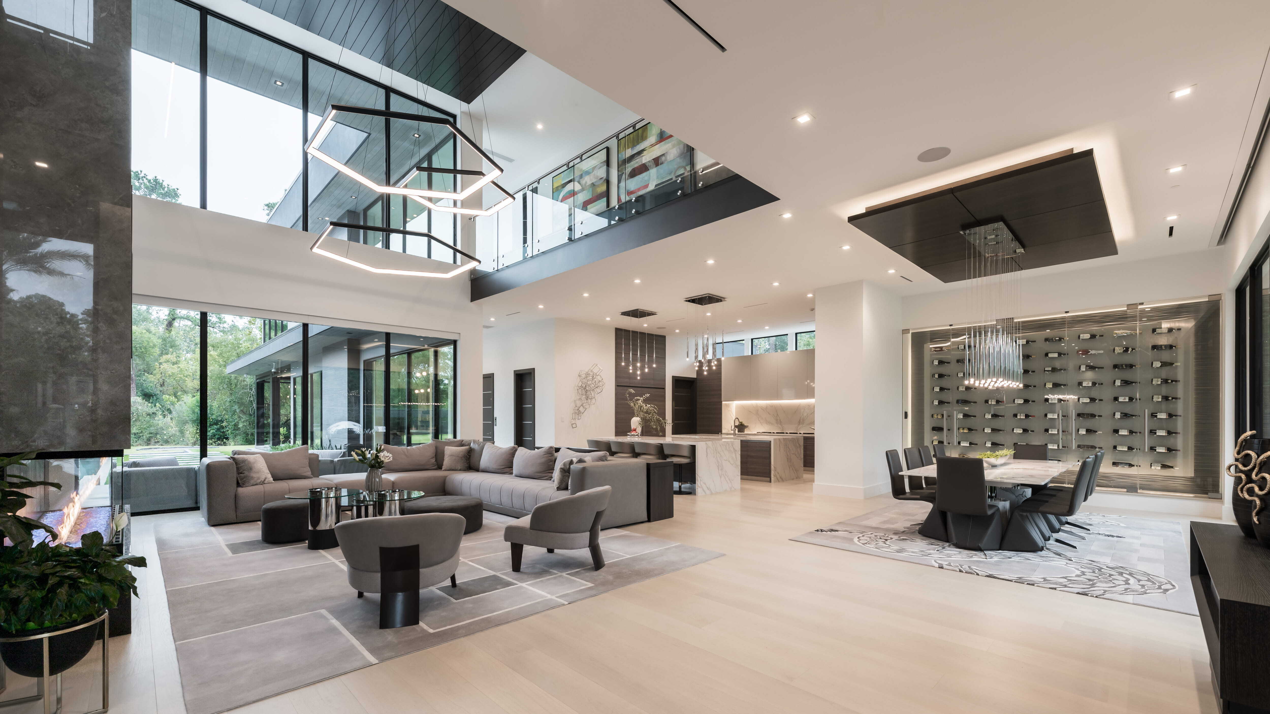 Open-concept living and dining space with two-story glass walls, modern fireplace, geometric chandelier, marble dining table, and a glass wine room in a contemporary Houston custom home.