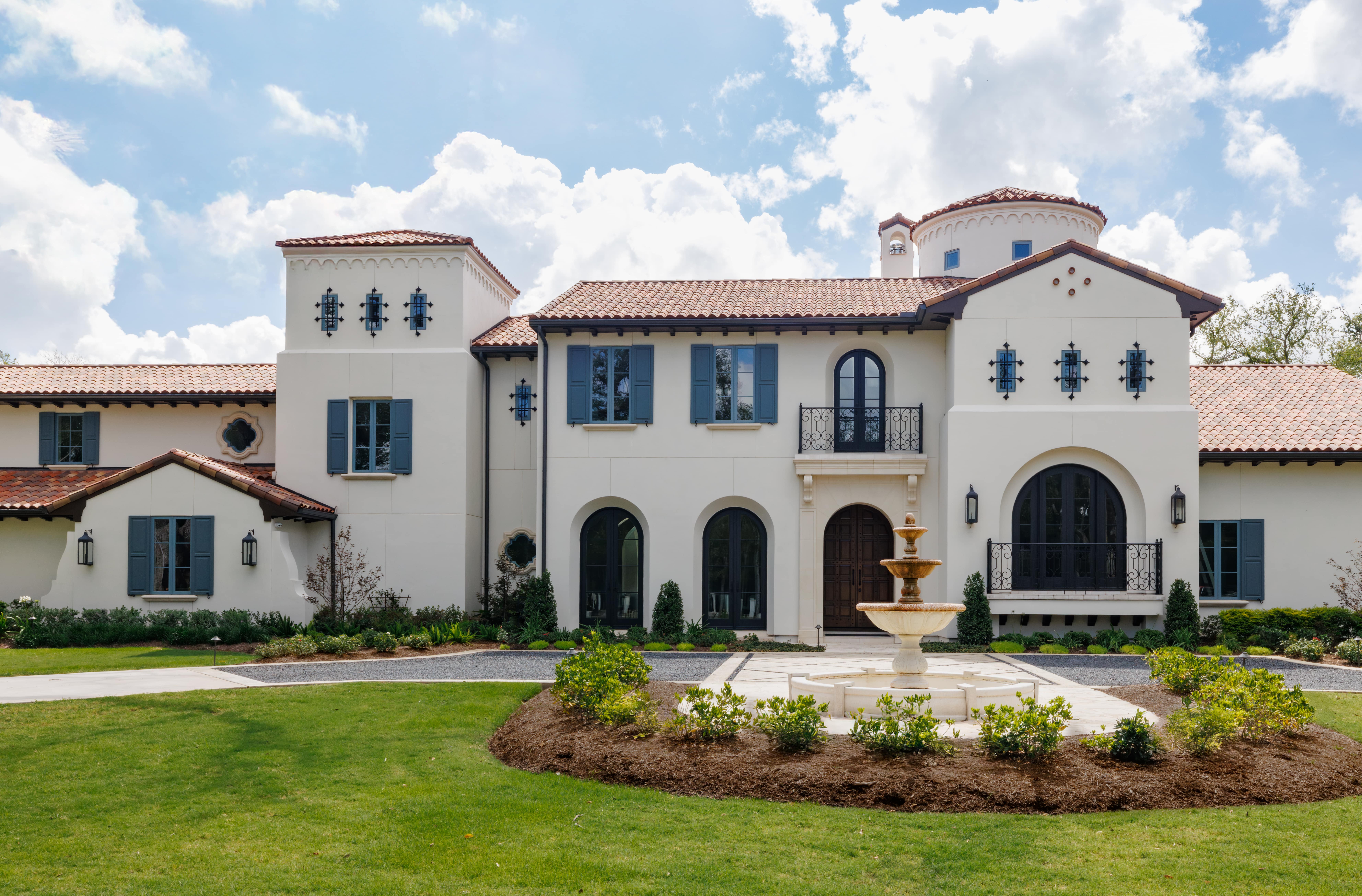 Sims Luxury Builders – Spanish Hacienda – Grand front elevation with arched windows, blue shutters, clay tile roof, wrought-iron balconies, and a central courtyard fountain.