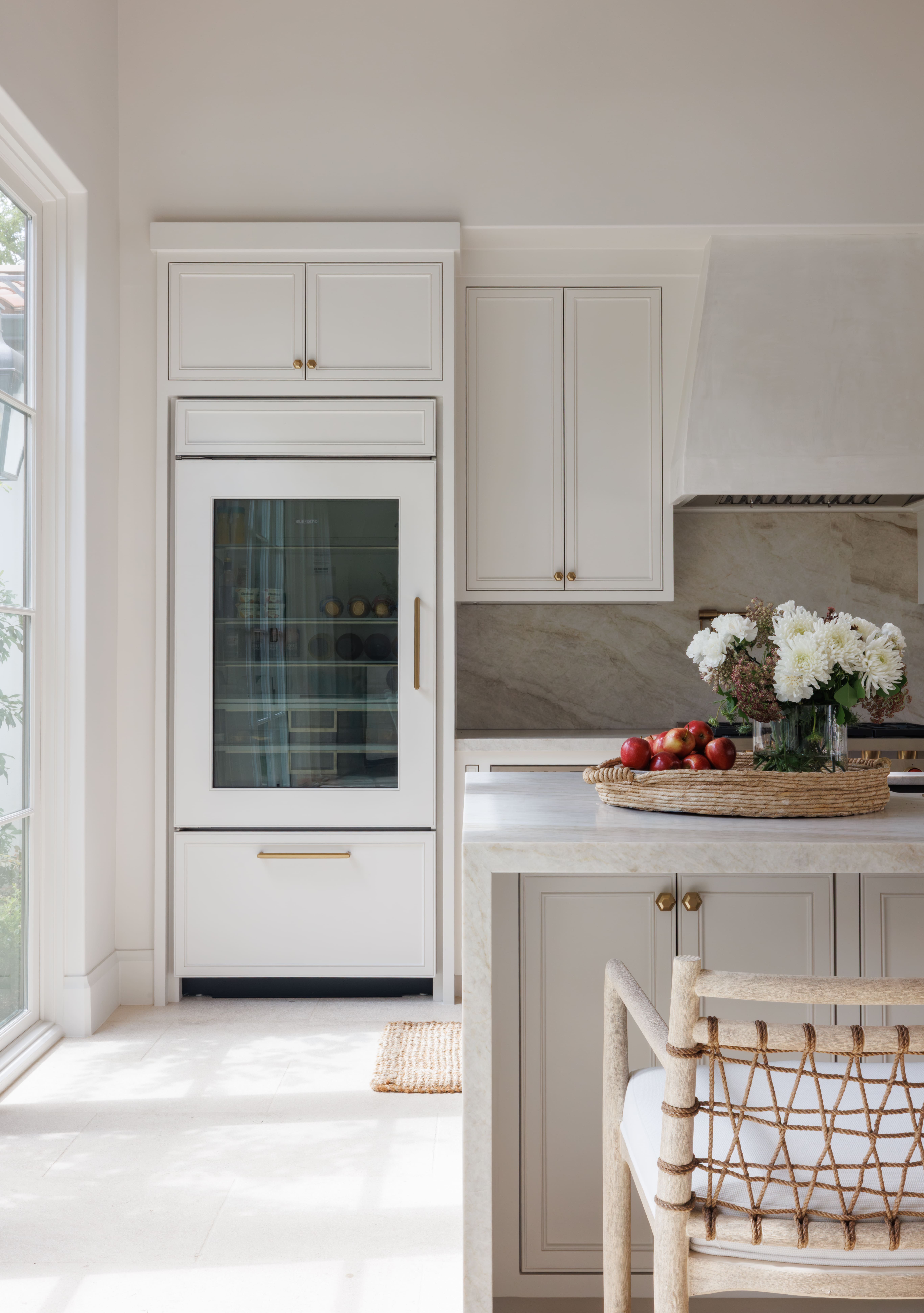 Spanish Hacienda – Bright kitchen detail featuring a paneled refrigerator with glass door, marble countertops, brass hardware, and a woven tray with flowers and apples.