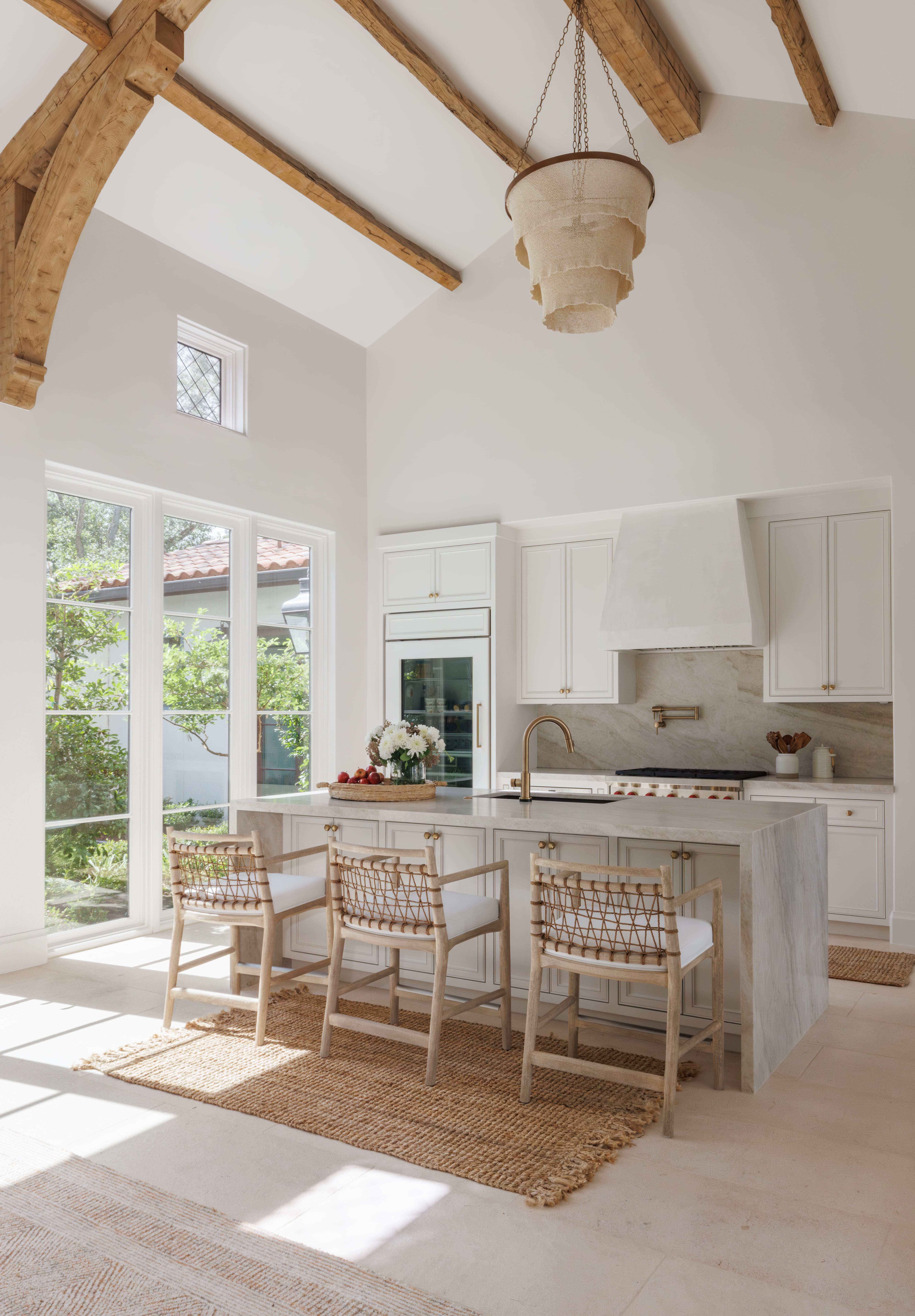 Spanish Hacienda - kitchen with vaulted ceiling and exposed wood trusses, white cabinetry, marble island, brass fixtures, and woven counter stools.