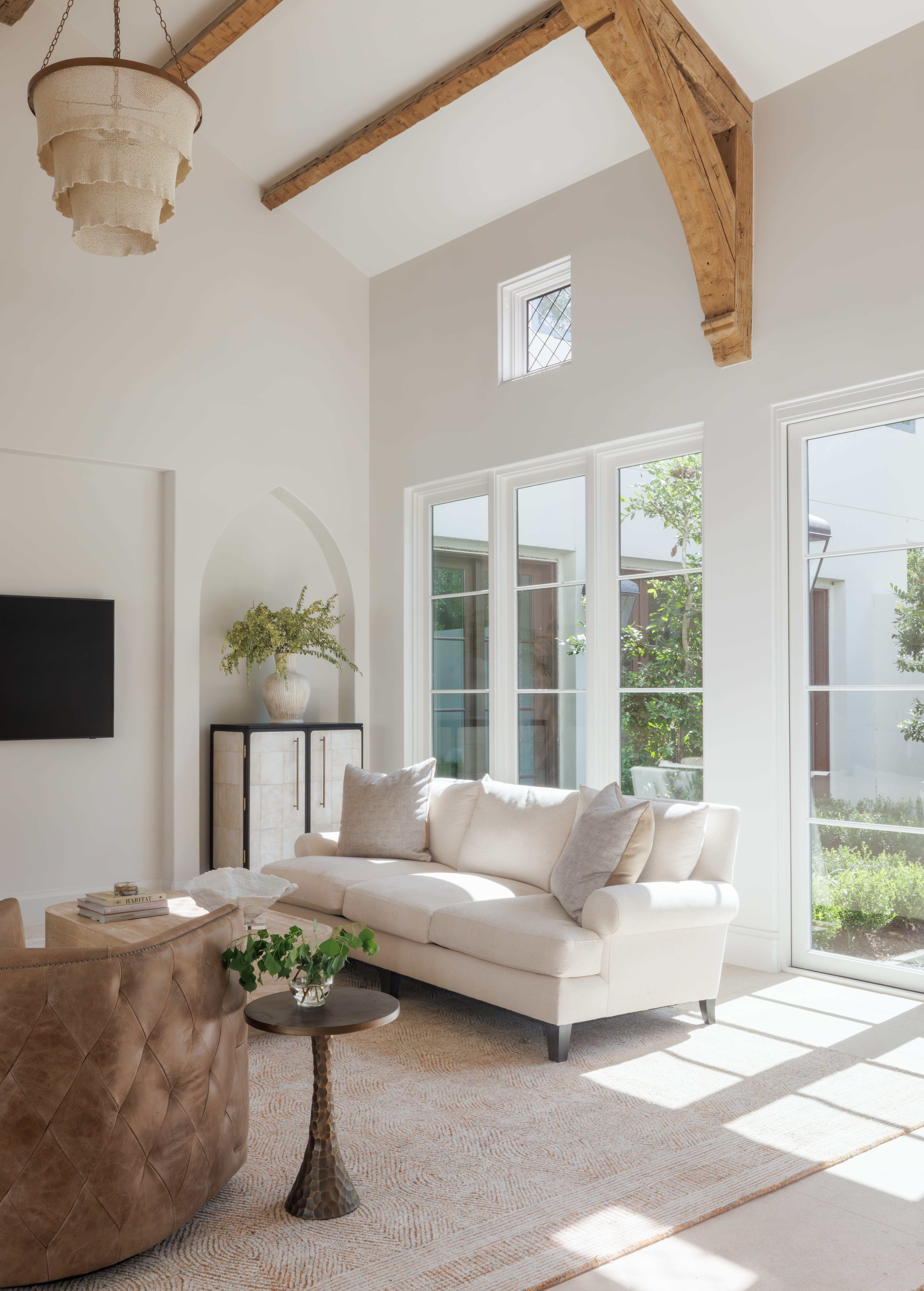 Spanish Hacienda - living room with cathedral ceiling, exposed beams, large windows, neutral sofa, leather swivel chair, and soft natural light filling the space.