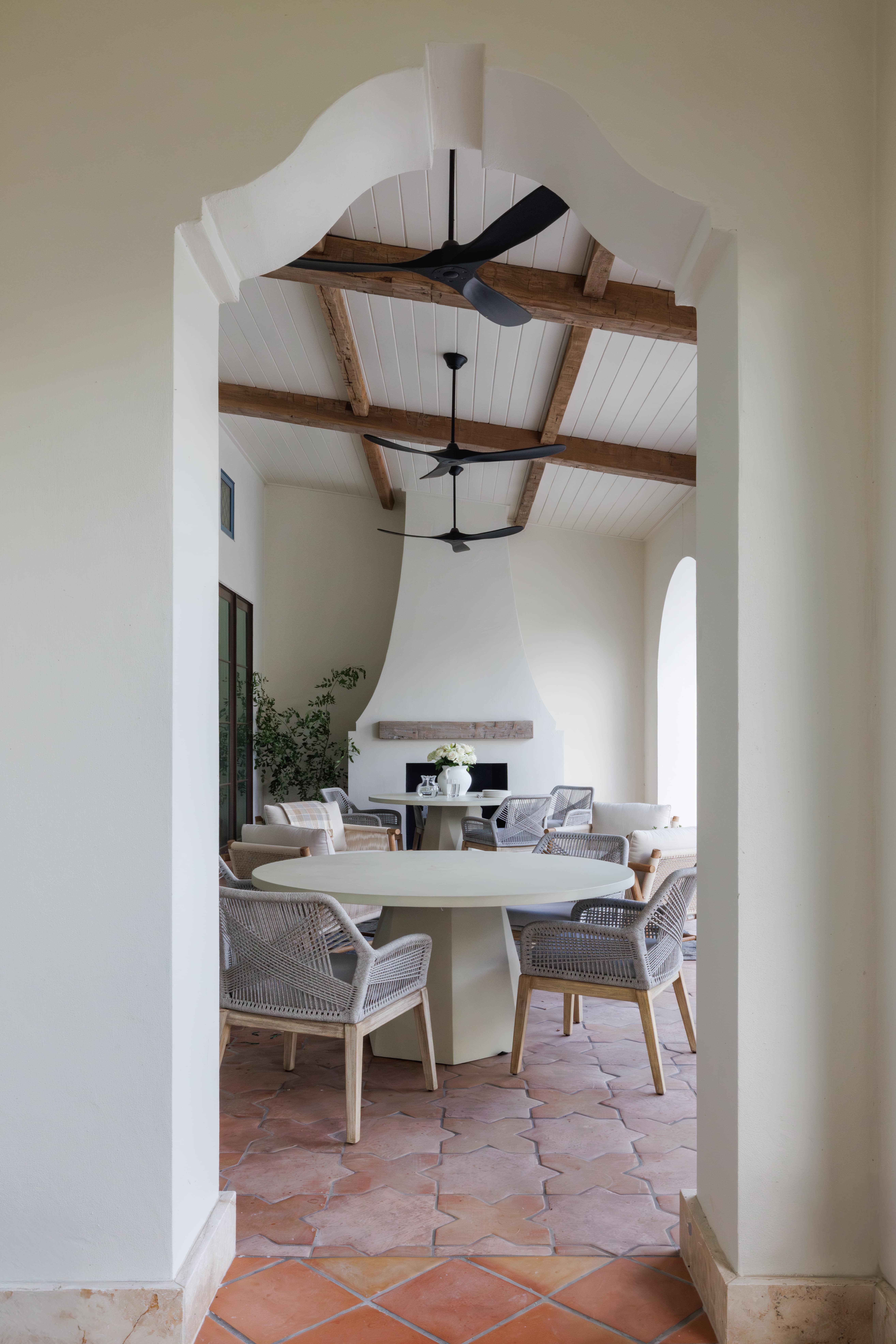 Spanish Hacienda - covered patio dining area framed by a decorative stucco arch, showing a round dining table, woven chairs, and wood-beam ceiling fans.