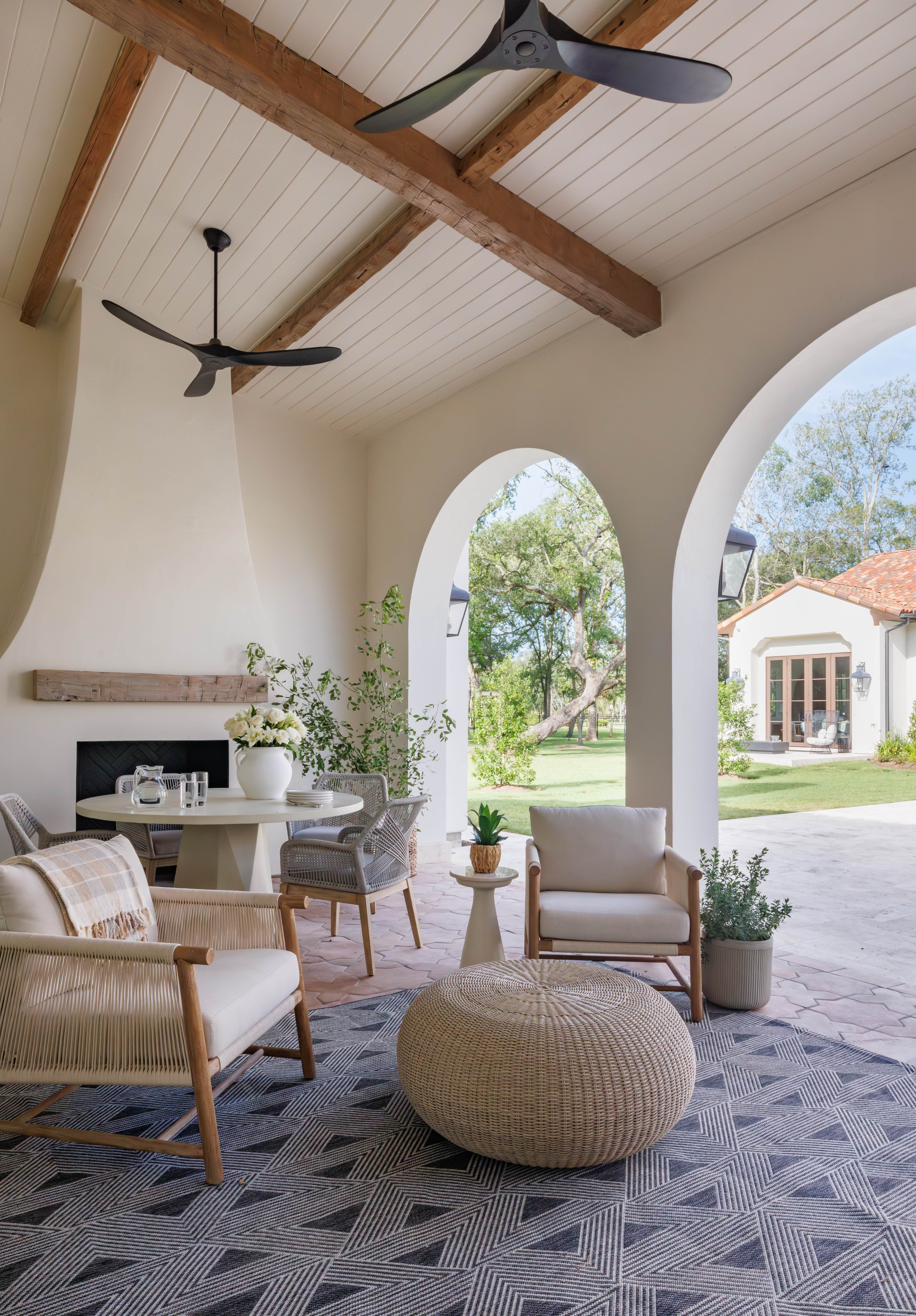 Spanish Hacienda - covered outdoor living space with arched openings, wood-beam ceiling, modern black fans, woven lounge chairs, round wicker ottoman, and a patterned rug overlooking the landscaped backyard.