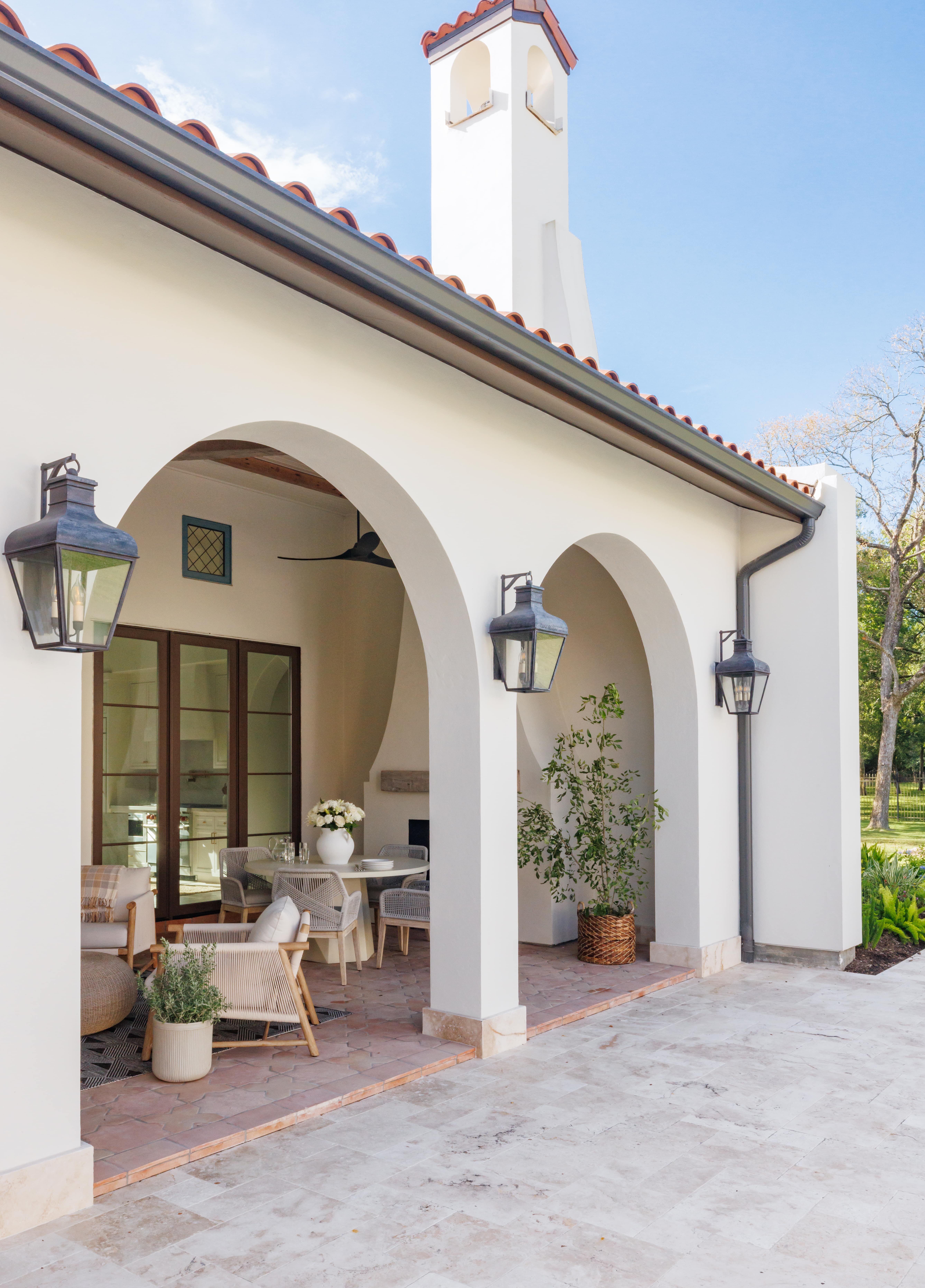 Spanish Hacienda - exterior patio with white stucco arches, clay tile roof, lantern sconces, and a covered seating area visible from the stone terrace.