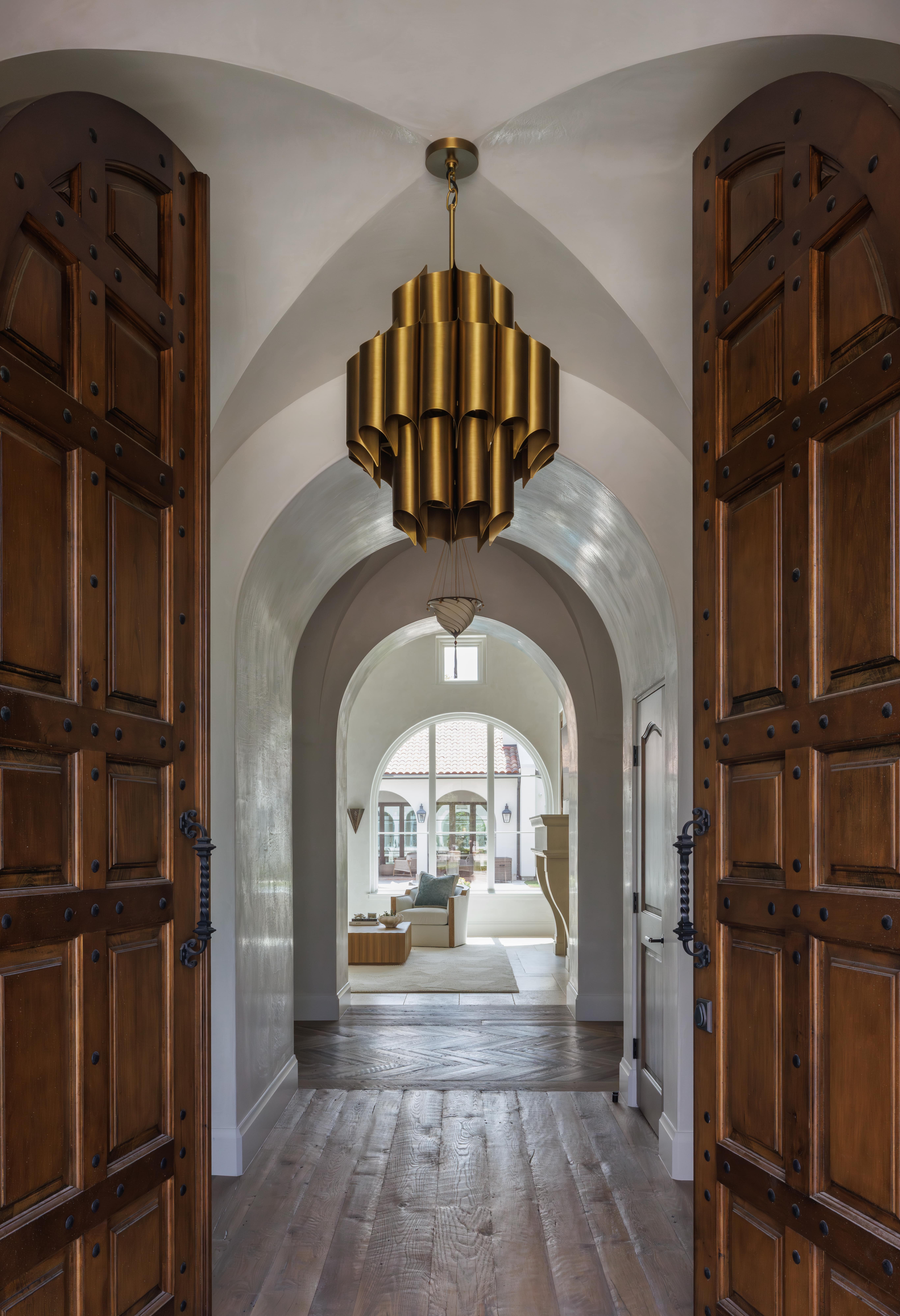Spanish Hacienda - entry hall with carved wooden doors, arched plaster ceiling, gold tiered chandelier, and a view through to the sunlit sitting room beyond.
