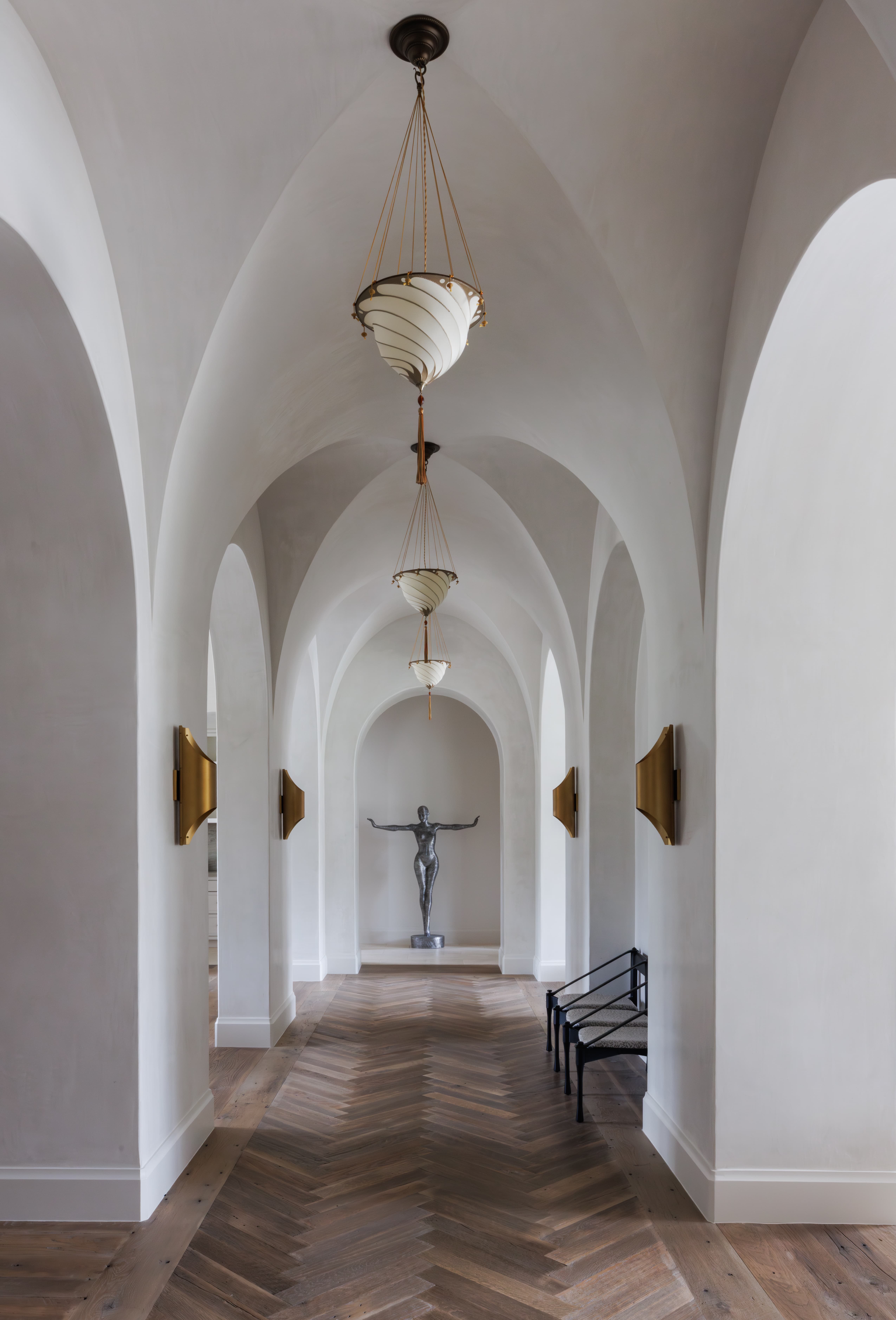 Spanish Hacienda - interior gallery hallway featuring vaulted plaster arches, brass lighting, chevron wood flooring, and a sculptural art niche at the end of the corridor.