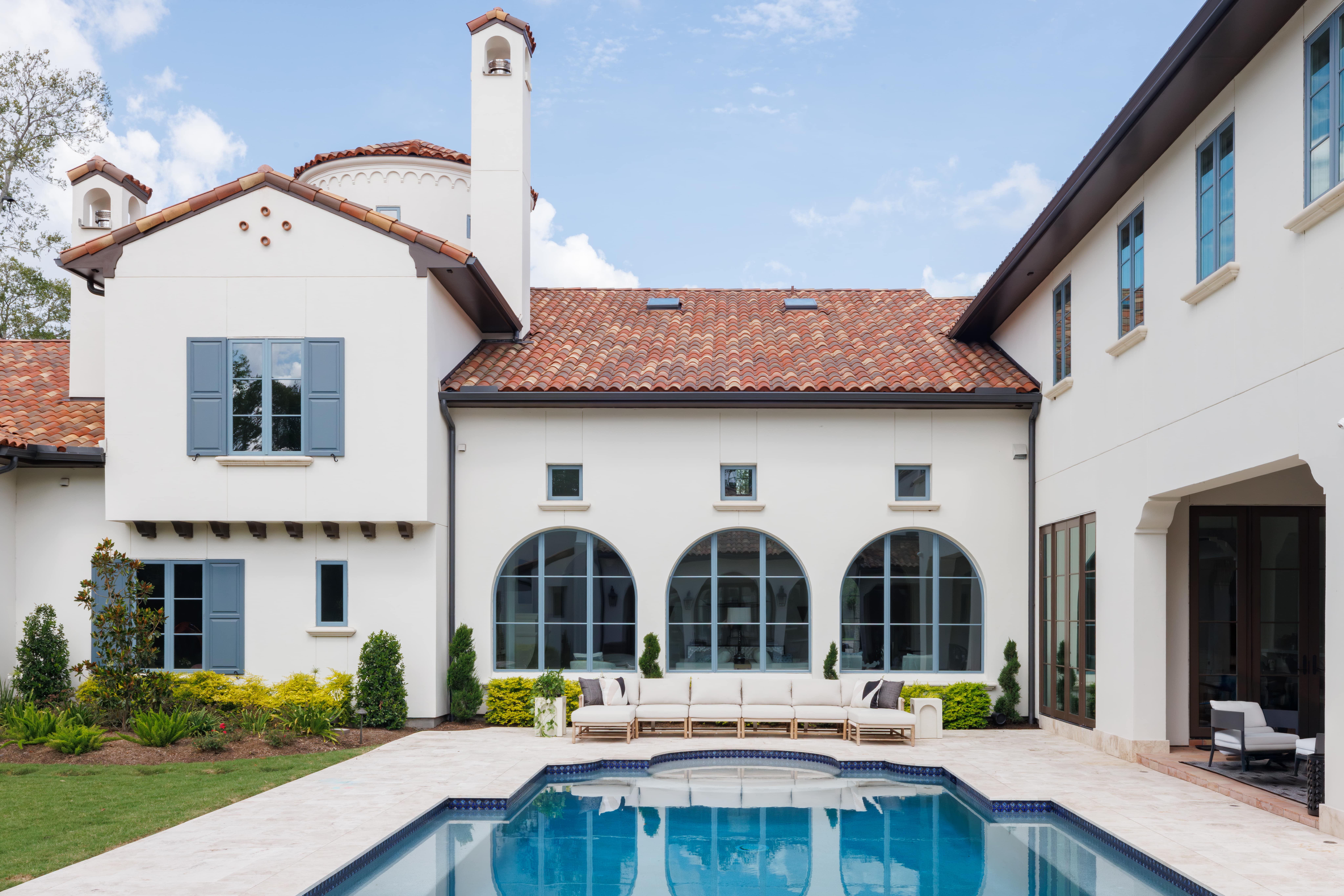 Sims Luxury Builders – Spanish Hacienda – Pool courtyard framed by arched windows, clay tile rooflines, blue shutters, and a long outdoor sofa along the rear façade.