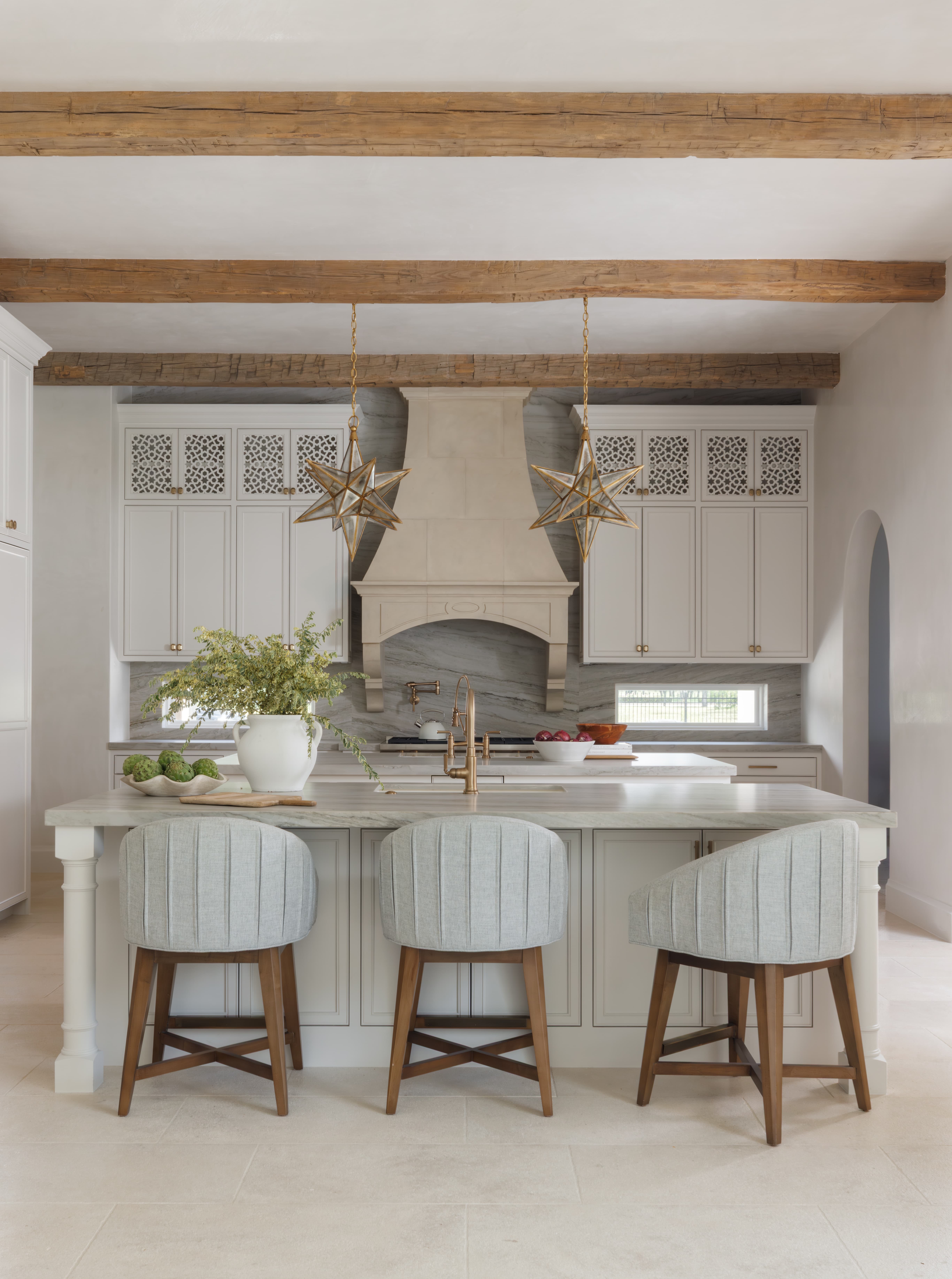 Spanish Hacienda - kitchen featuring star-shaped pendant lights, marble island with upholstered barstools, plaster range hood, and carved upper cabinet panels beneath rustic ceiling beams.