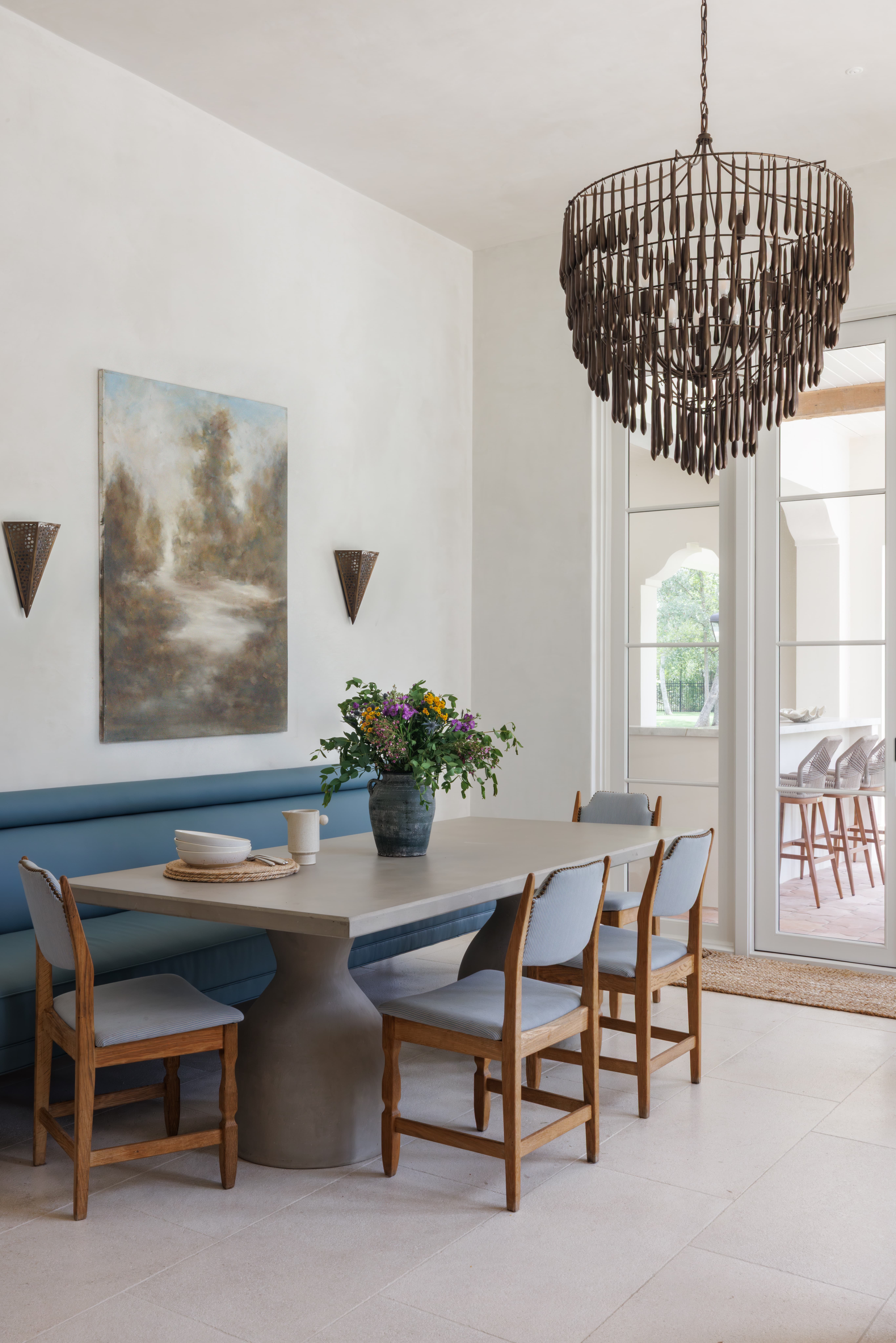 Spanish Hacienda - breakfast nook with sculptural concrete table, blue upholstered banquette, wood dining chairs, and a bronze cascading chandelier beside French doors.