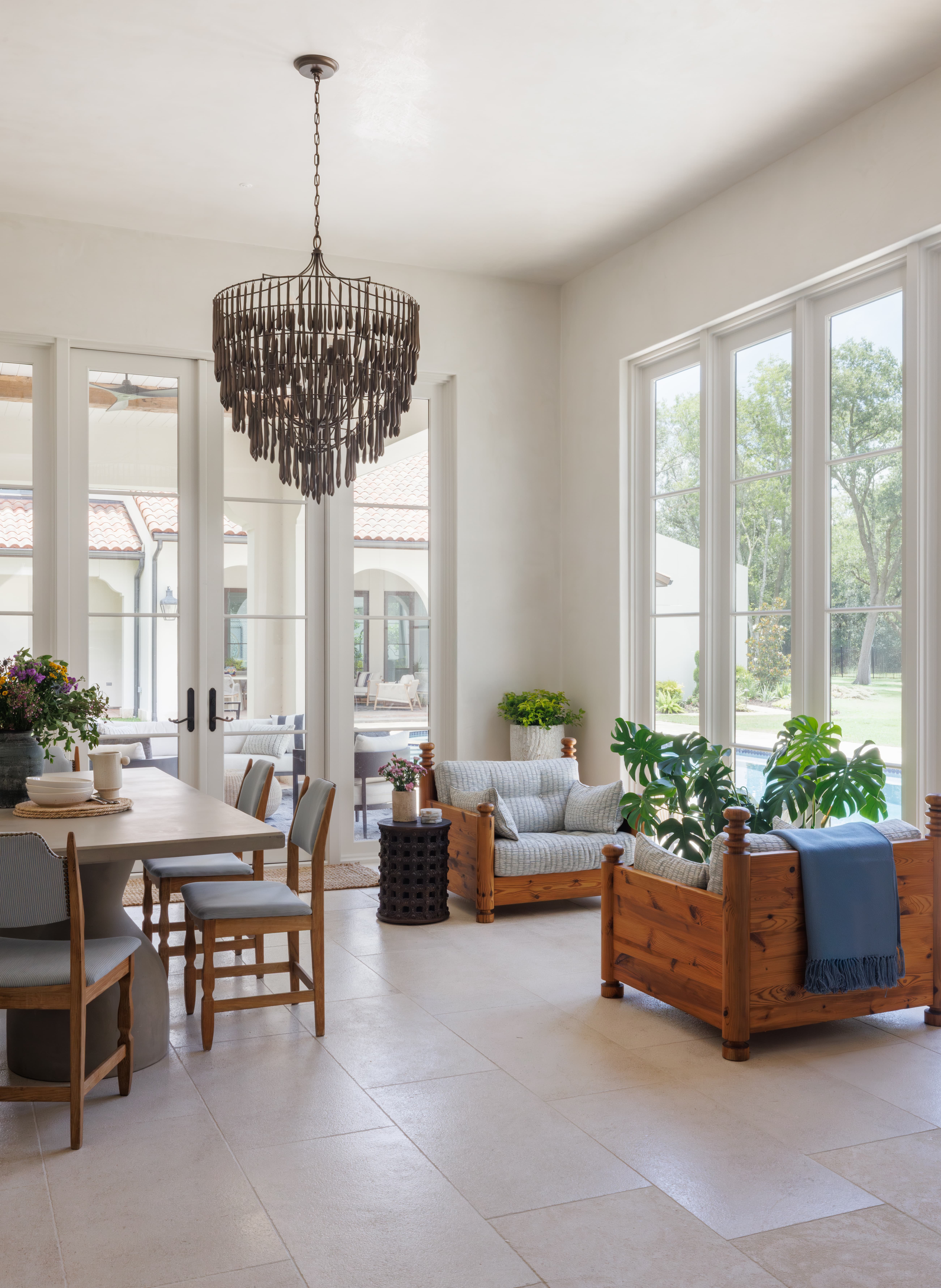 Spanish Hacienda - sunroom with tiered bronze chandelier, light stone flooring, warm wood lounge seating, and full-height windows overlooking the pool.