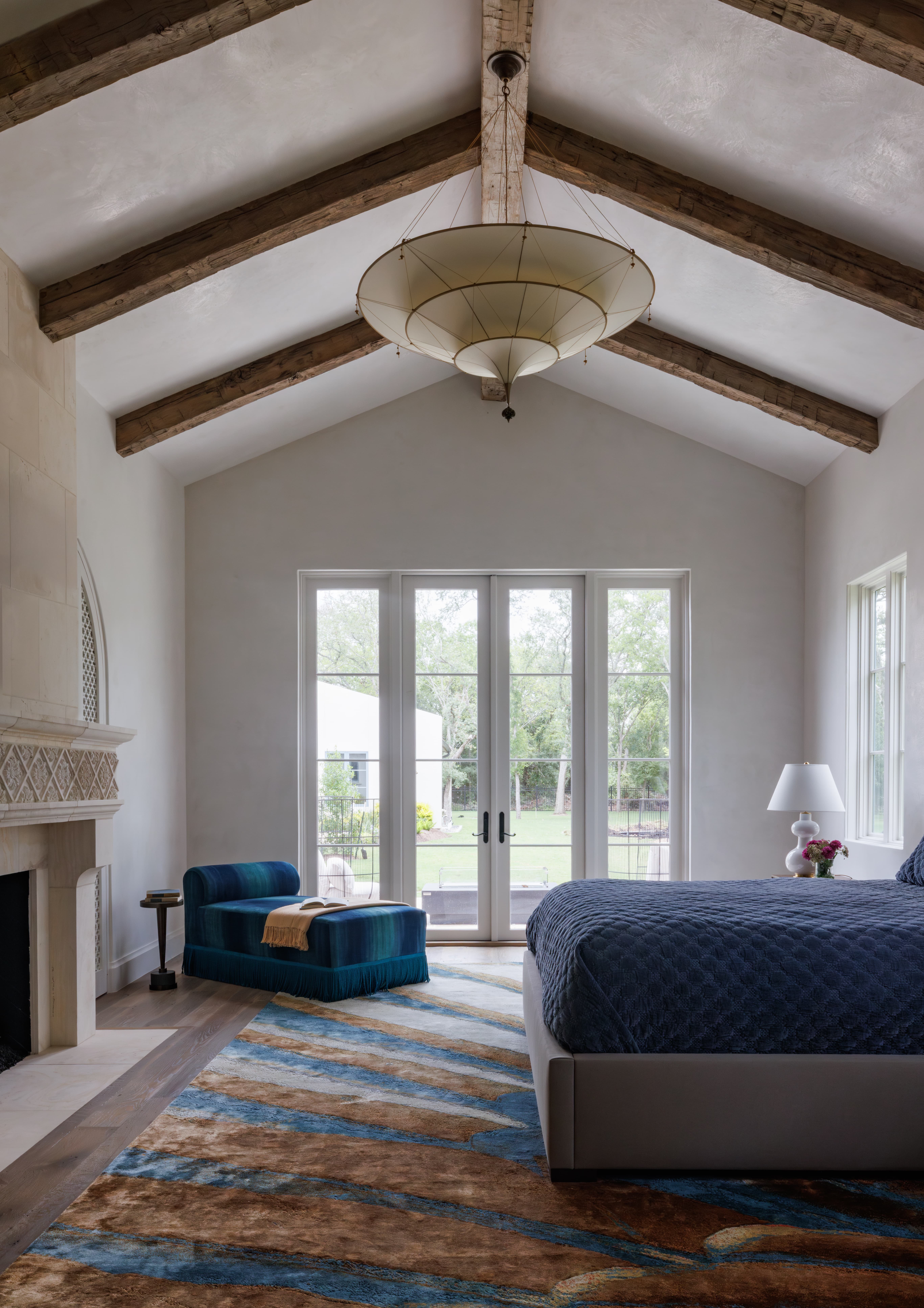 Spanish Hacienda - bedroom with vaulted ceiling, exposed wood beams, large sculptural pendant light, blue chaise lounge, patterned area rug, and French doors opening to the backyard.
