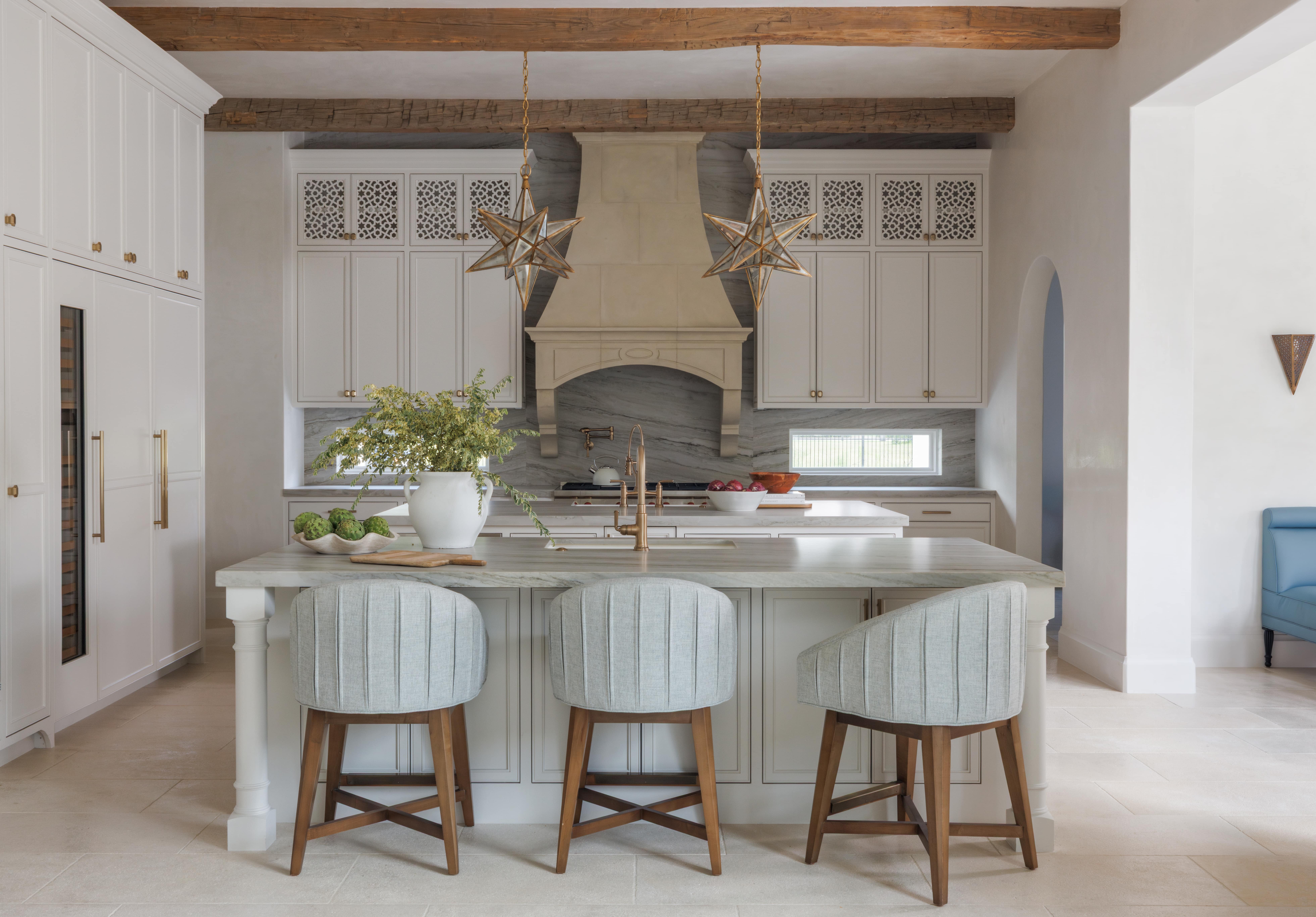 Spanish Hacienda – Elegant kitchen with carved stone range hood, star-shaped brass pendants, marble countertops, and upholstered counter stools beneath wood ceiling beams.