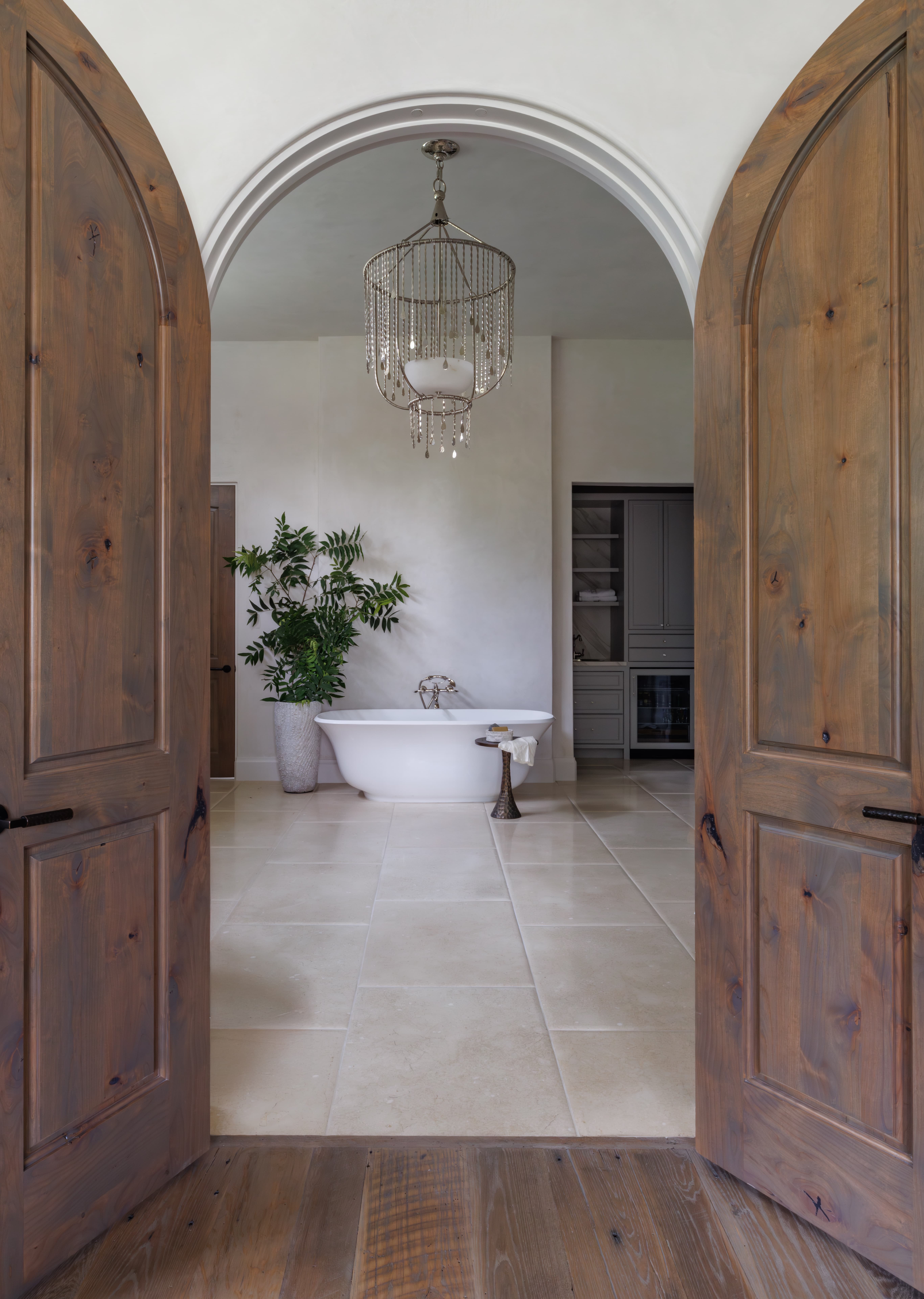 Spanish Hacienda – Arched wooden double doors opening to a serene bathroom with a freestanding tub, crystal chandelier, large potted greenery, and warm stone flooring.