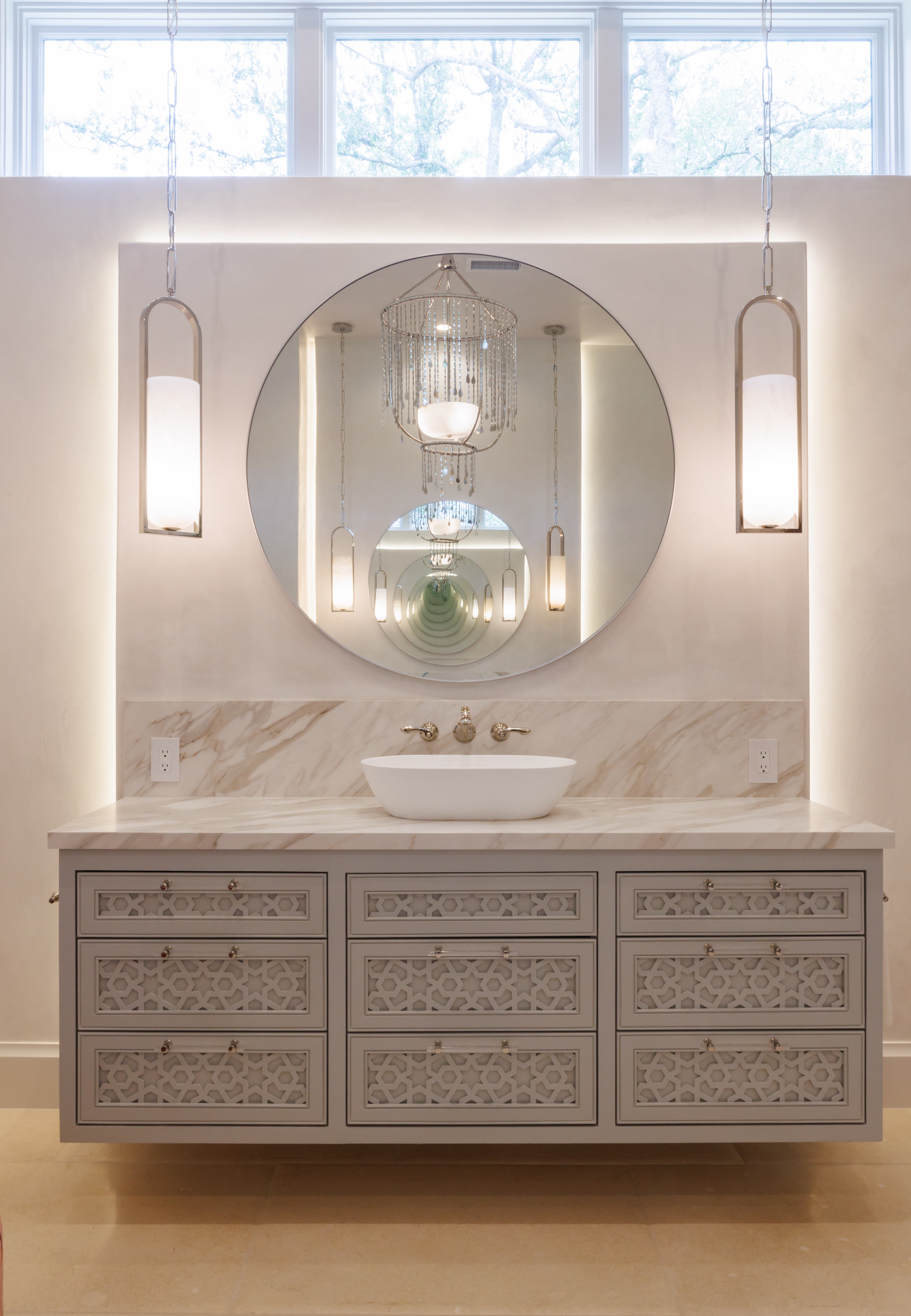 Spanish Hacienda – Floating bathroom vanity with carved drawer fronts, vessel sink, illuminated round mirror, and pendant sconces reflecting a crystal chandelier.