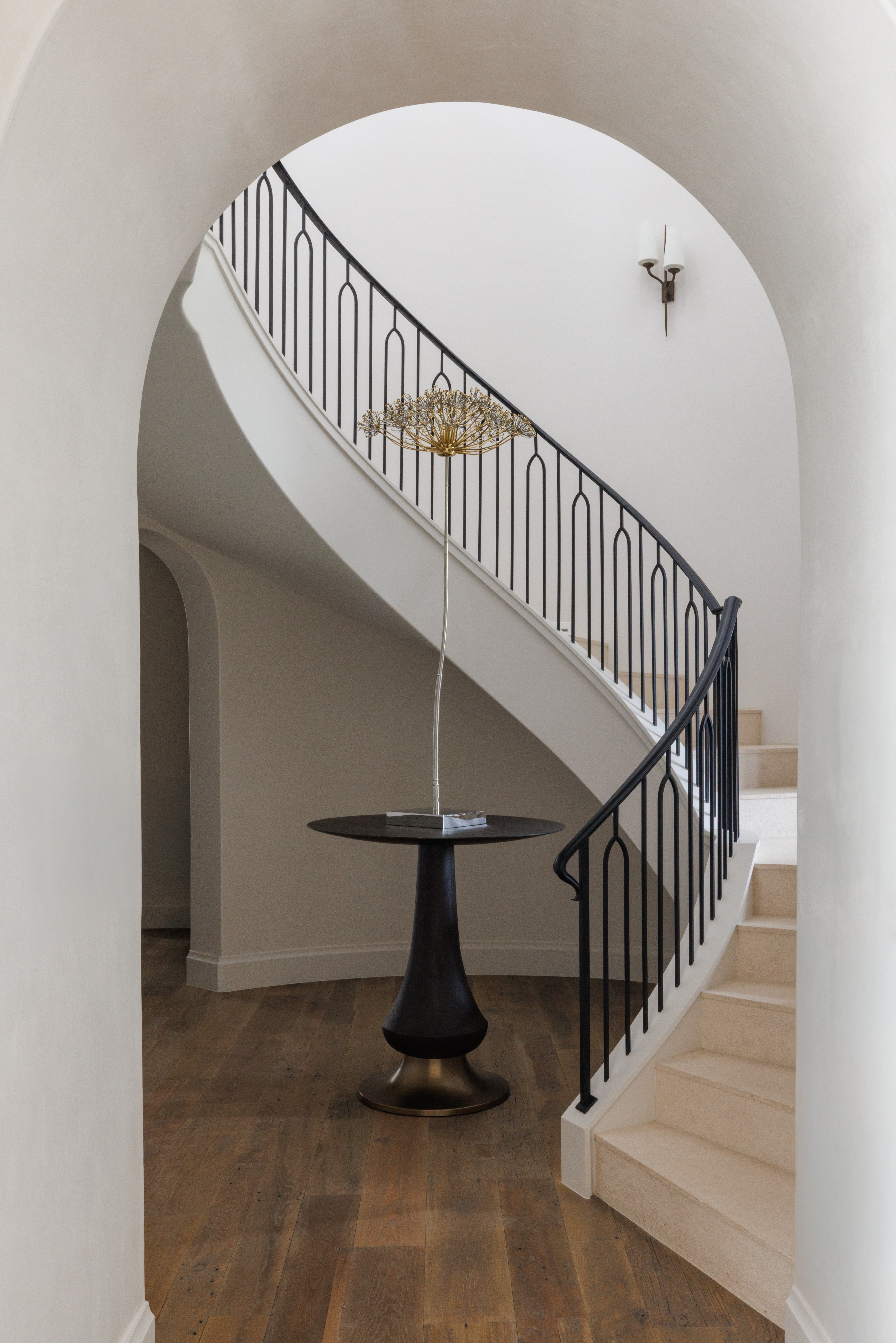 Spanish Hacienda – Curved staircase framed by an arched entry, featuring iron railings, wide plank wood flooring, and a sculptural accent table.