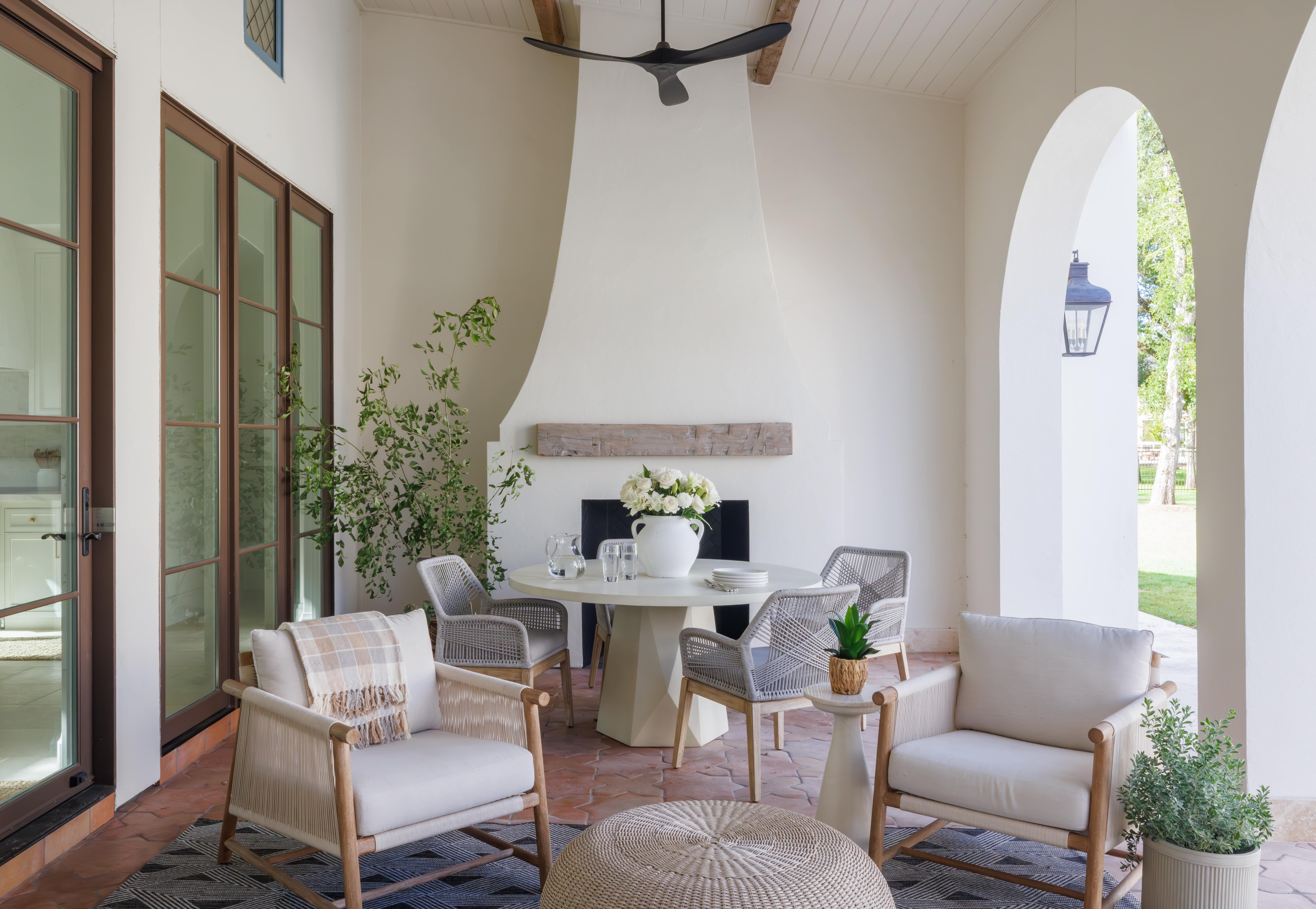 Spanish Hacienda – Covered outdoor loggia with smooth plaster fireplace, woven chairs, round dining table, and arched openings overlooking the lawn.