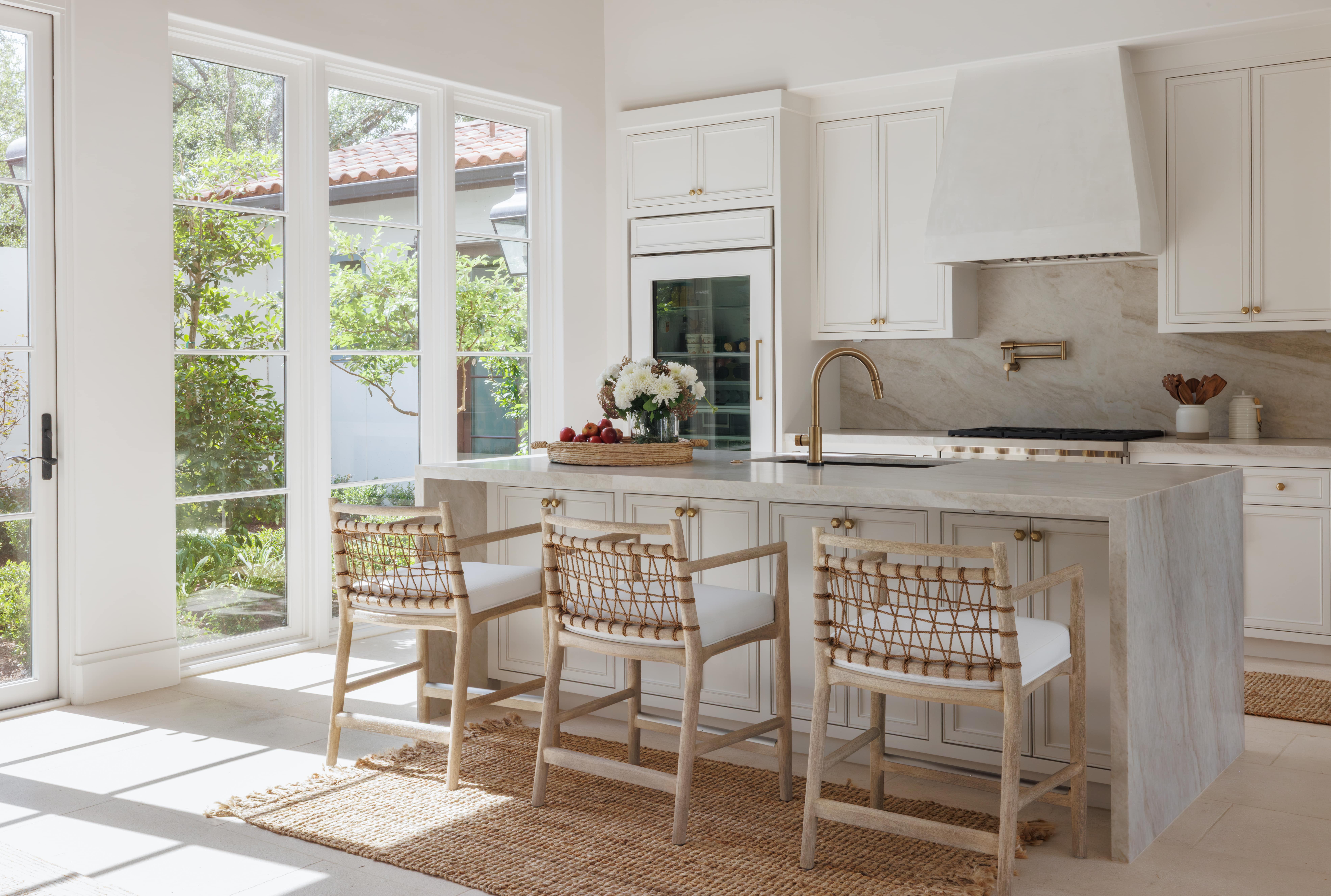 Spanish Hacienda – Sunlit kitchen with floor-to-ceiling windows, marble waterfall island, woven barstools, and brass fixtures.
