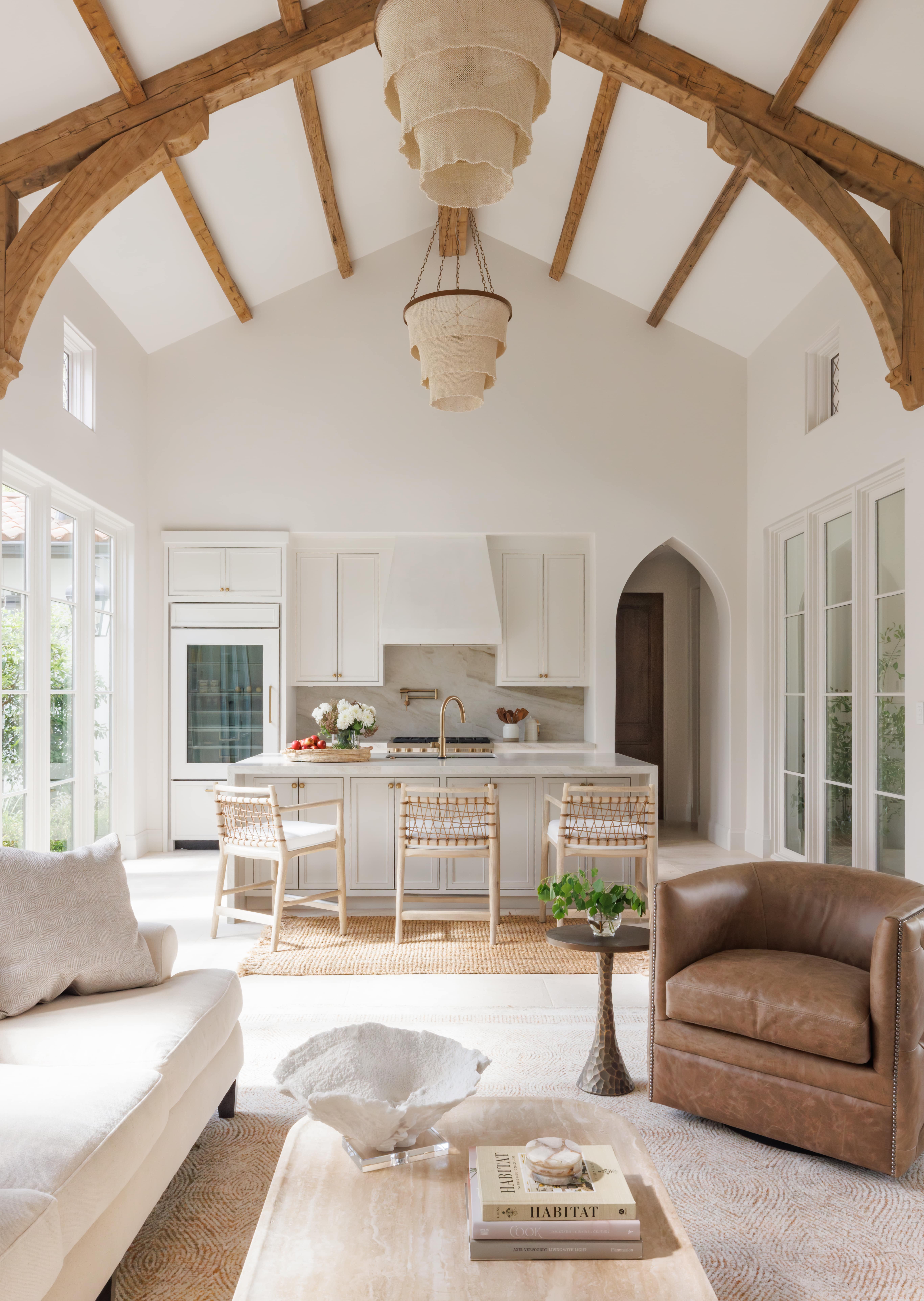 Spanish Hacienda – Open living and kitchen space with exposed timber trusses, marble island, woven barstools, soft neutral furnishings, and abundant natural light.
