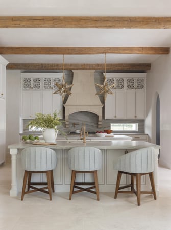 Sims Luxury Builders – Spanish Hacienda – Warm, inviting kitchen with star-shaped brass pendants, stone range hood, marble backsplash, light gray cabinetry, and upholstered counter stools under exposed wood ceiling beams.