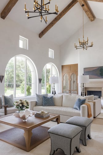 Sims Luxury Builders – Spanish Hacienda – Light-filled living room with arched windows, timber ceiling beams, iron chandeliers, cream sofa with blue accent pillows, carved wood doors, and a stone fireplace.