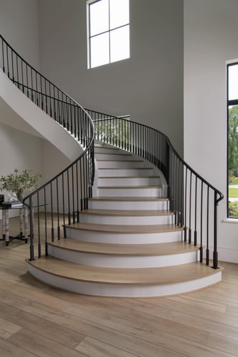 Curved staircase with light wood steps and black railings ascending through a bright, window-lit foyer.