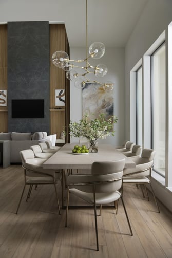 Dining area with a long stone table, upholstered chairs, brass chandelier, and artwork beside a tall dark fireplace wall.