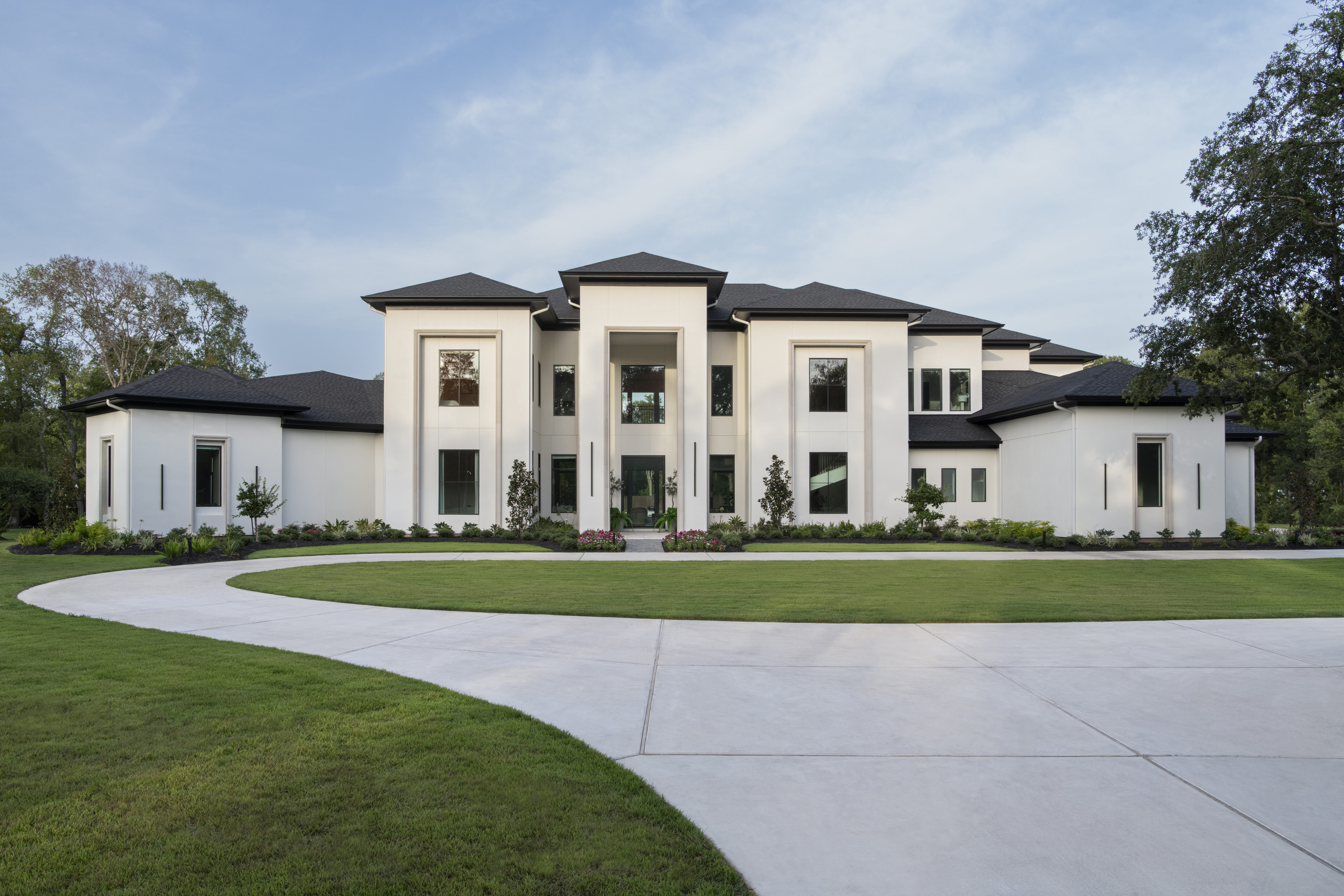 Front elevation of a modern transitional estate with white stucco, dark rooflines, and a circular driveway framed by manicured landscaping.