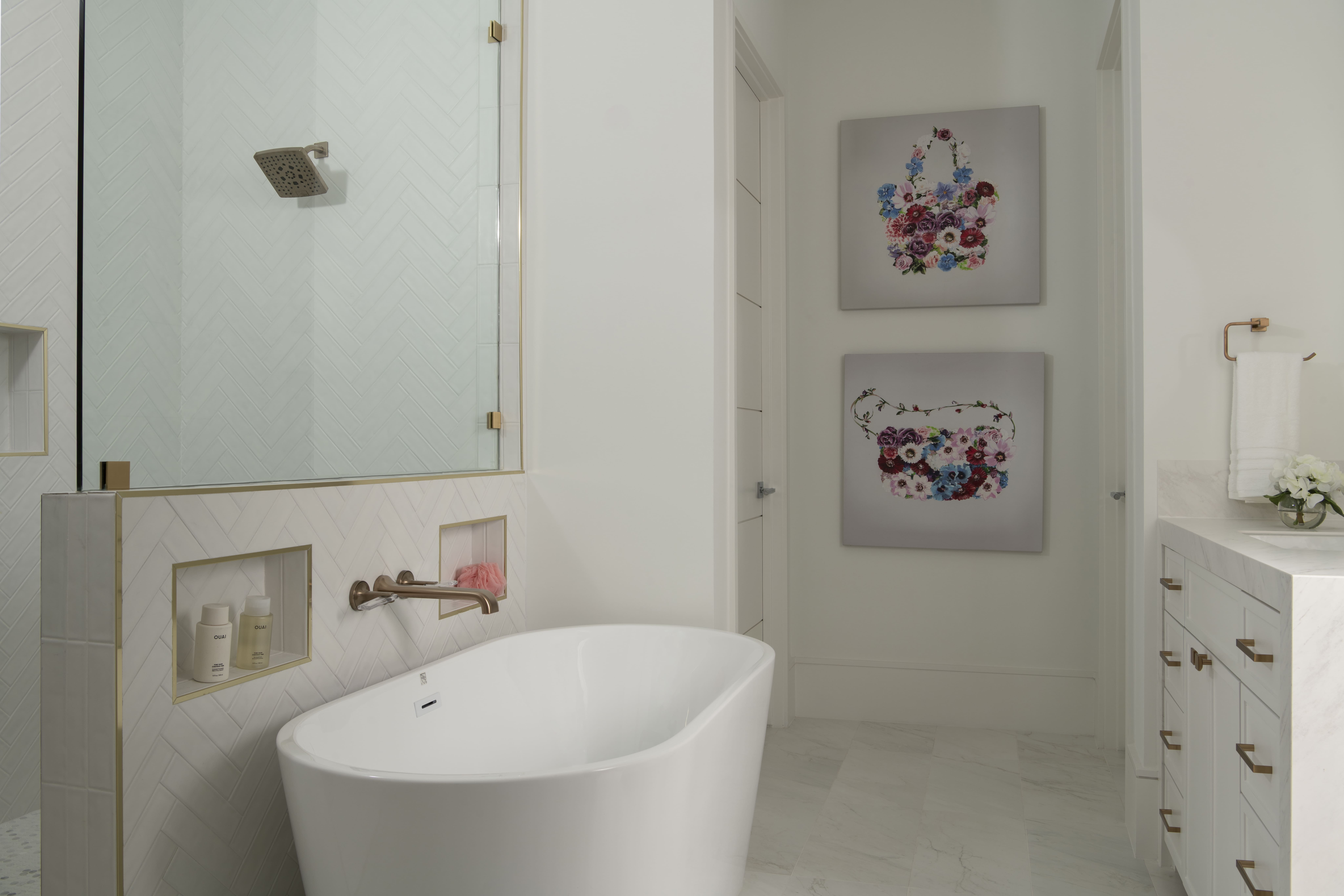 Bathroom with a freestanding white soaking tub, herringbone tile shower with brass fixtures, and floral artwork on the wall.