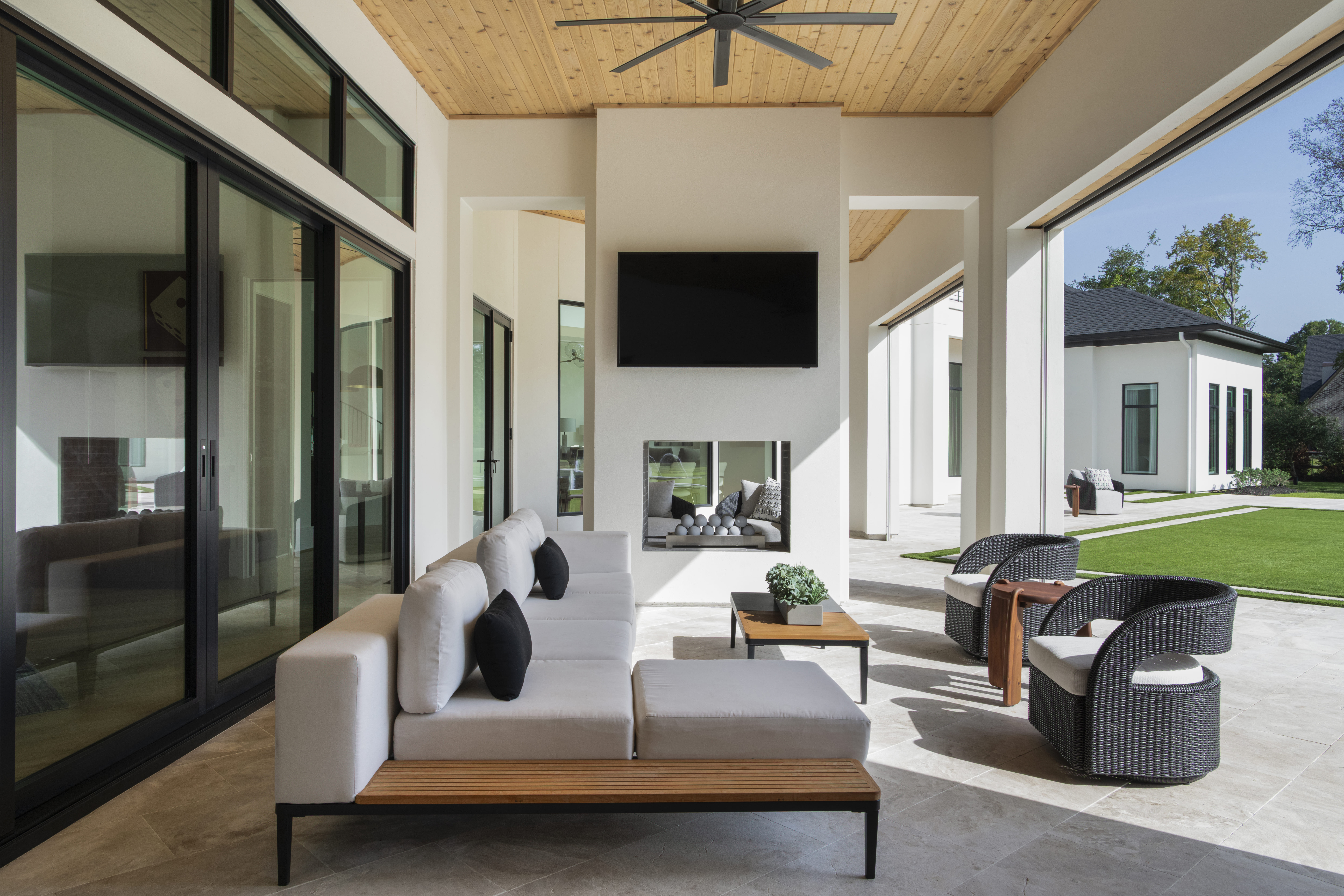 Covered outdoor living area with modern seating, large sliding glass doors, a TV over a sleek fireplace, and views of the lawn.