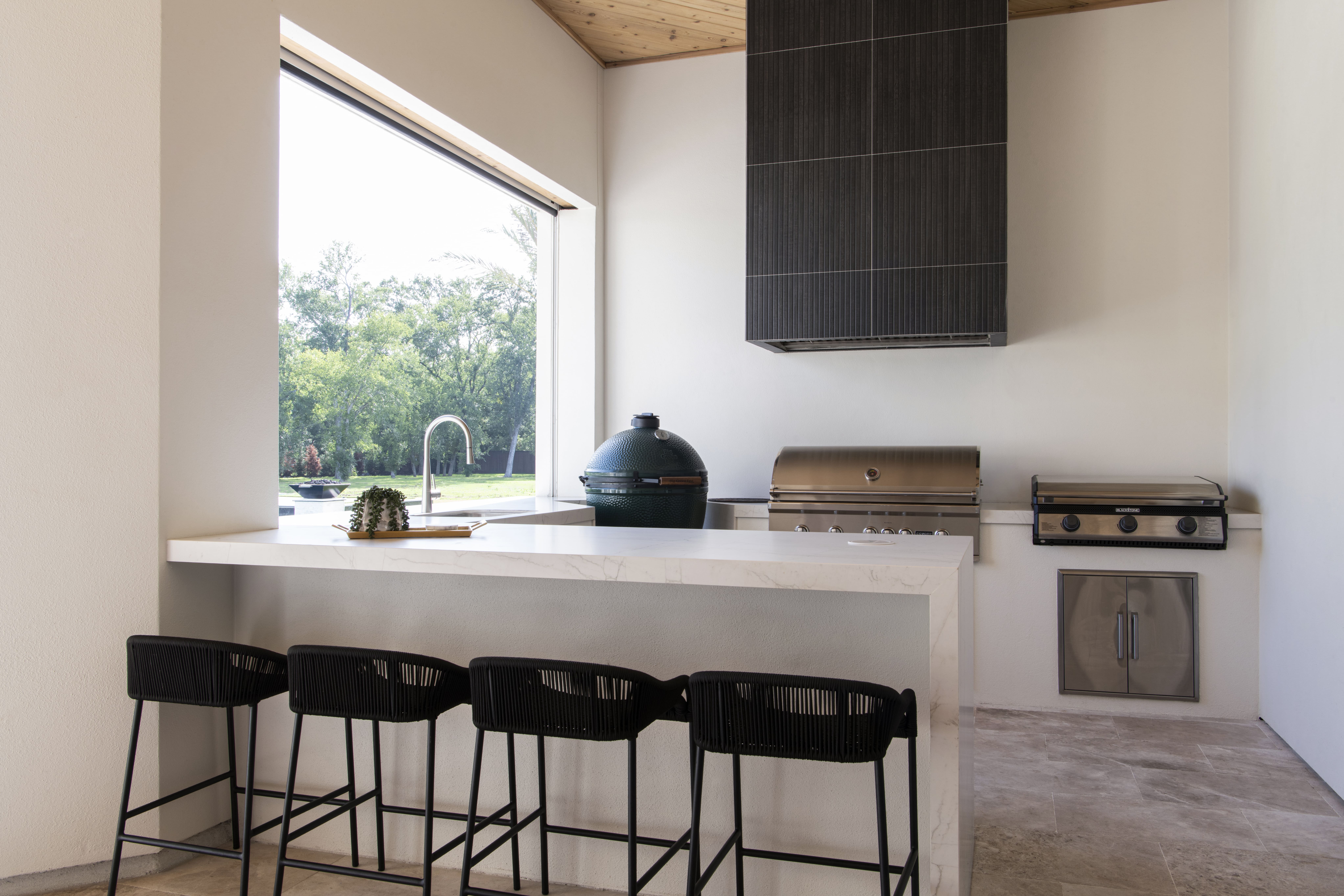 Outdoor kitchen with a marble island, black barstools, stainless grill, Big Green Egg smoker, and large passthrough window overlooking the lawn.